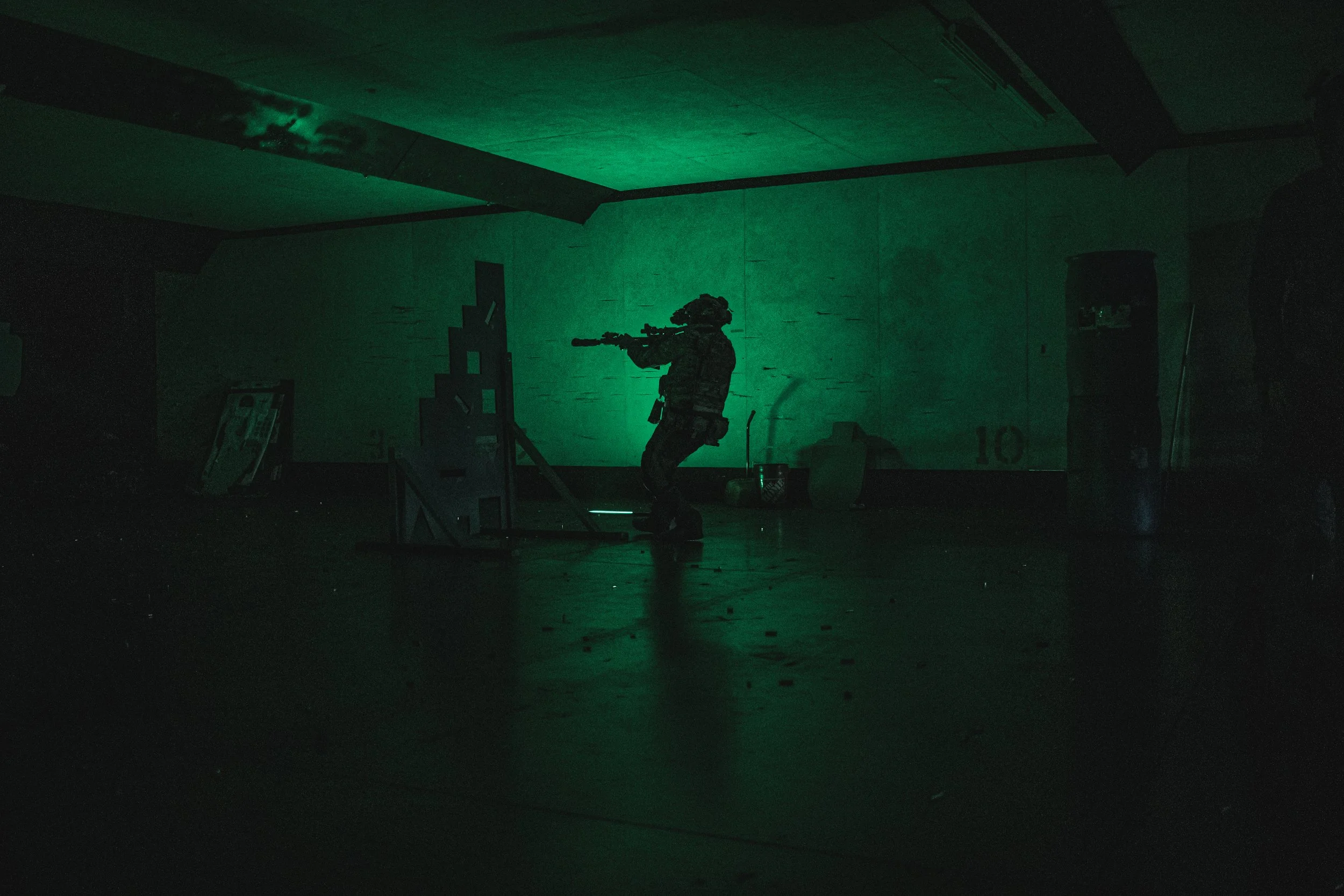 A soldier in tactical gear aiming a rifle while moving in a dark indoor training facility with green lighting.
