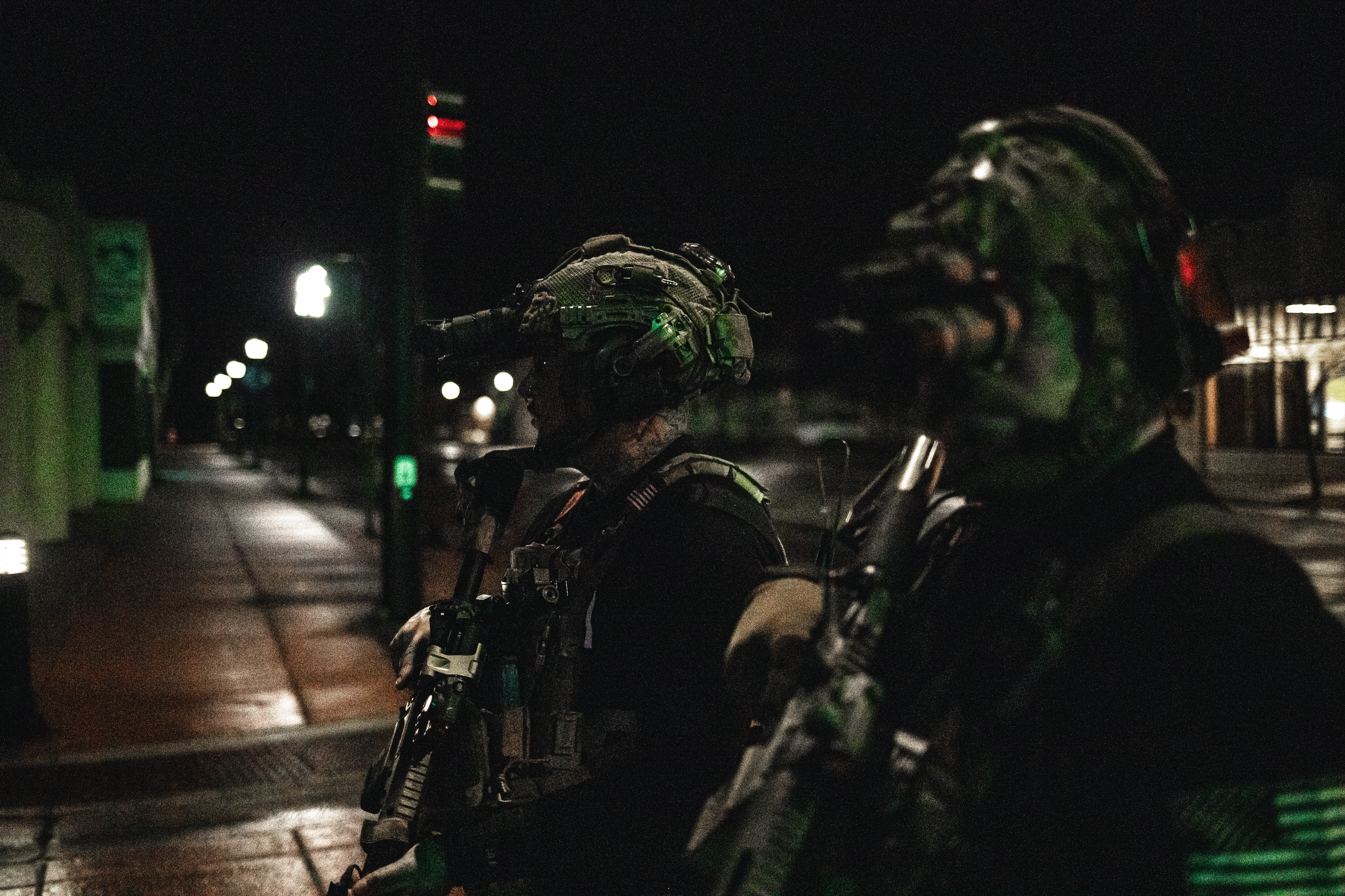 Two soldiers in full gear and helmets sitting on motorcycles in a dark, empty city street at night.