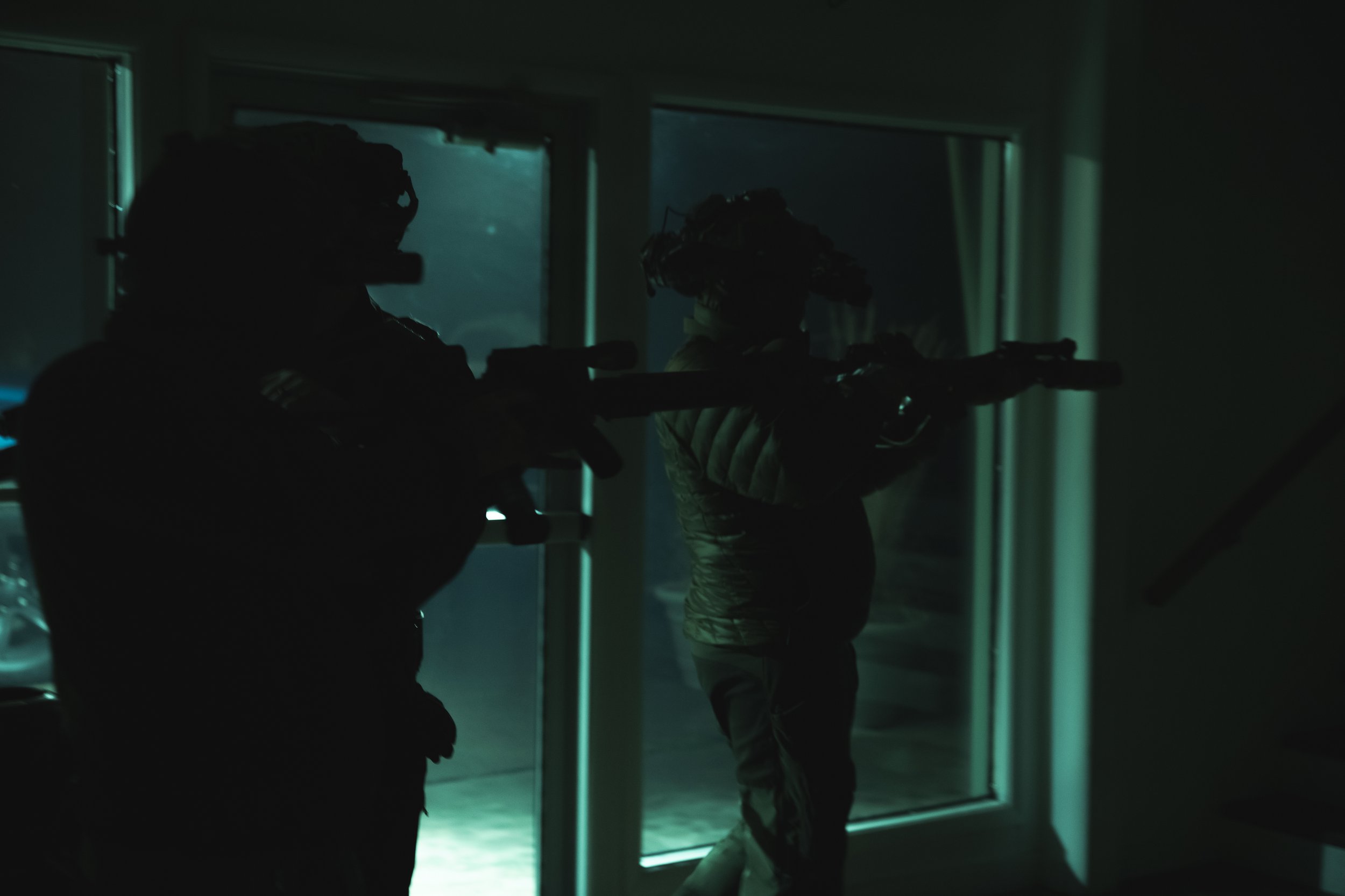 Two soldiers in tactical gear aiming rifles in a dark room with a glass door, backlit by dim teal light from outside.