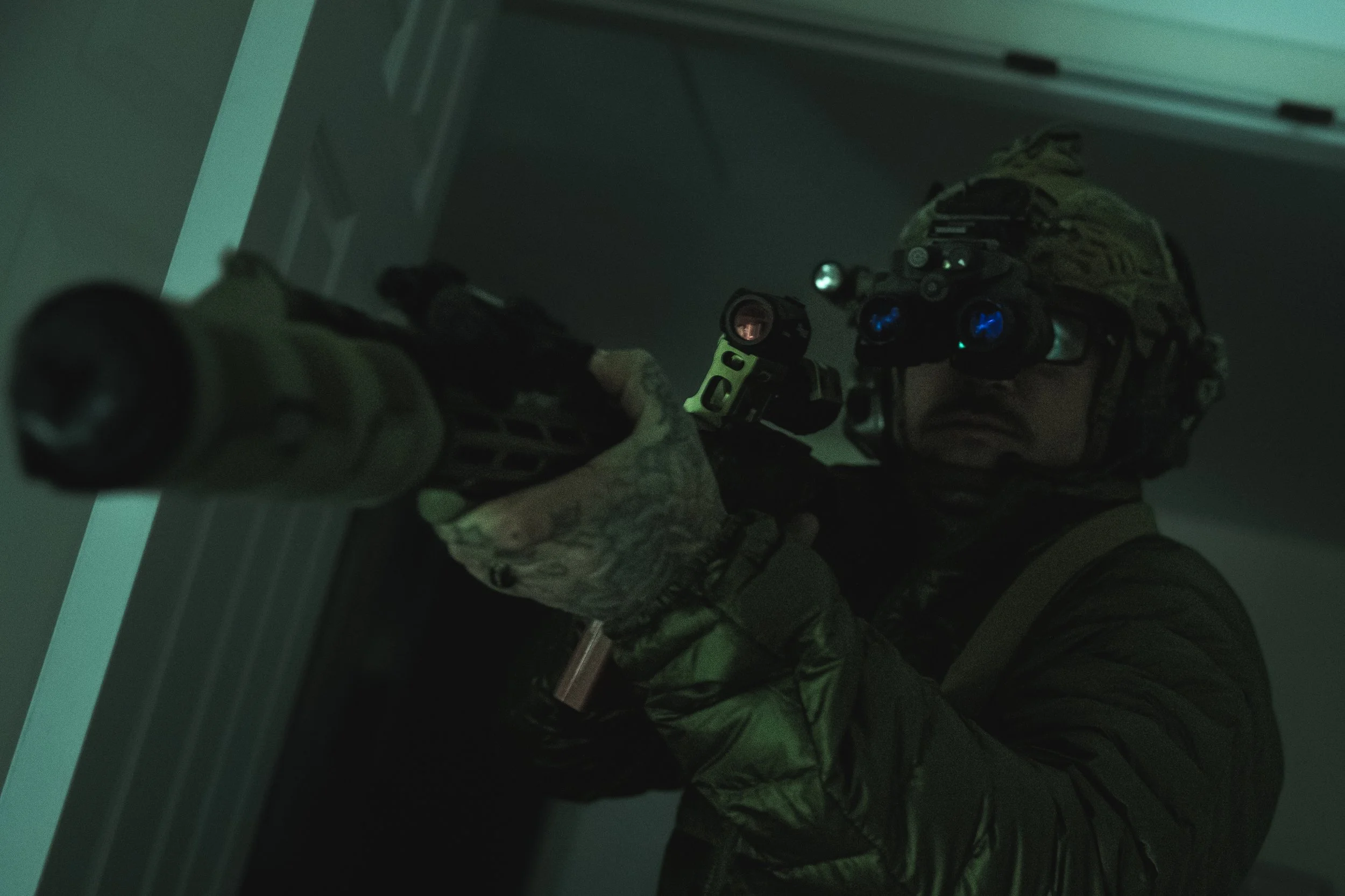 A soldier wearing night vision goggles aiming a firearm in a dimly lit room.