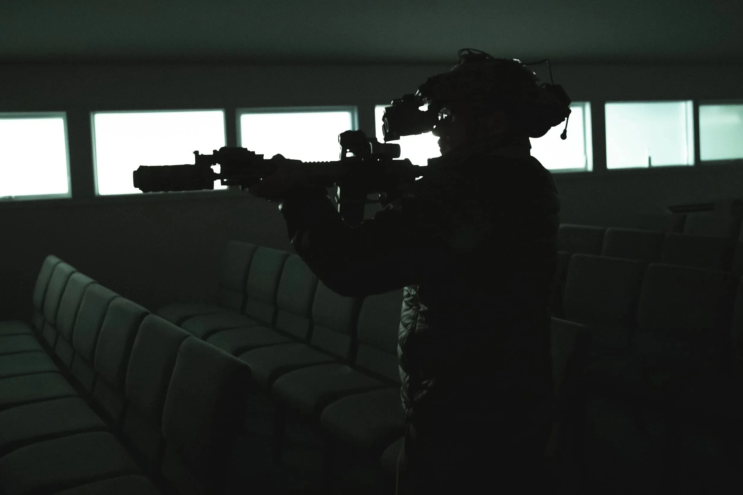 Silhouette of a soldier aiming a rifle in an indoor shooting range with windows in the background.