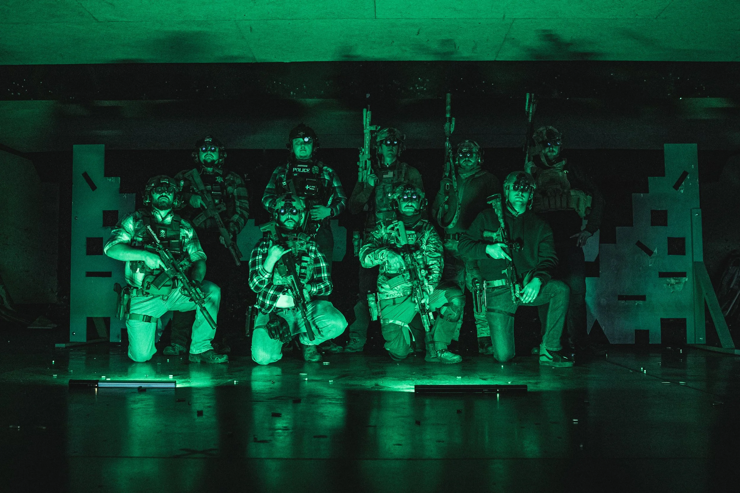 Group of eleven individuals in tactical gear holding rifles, posing for a photo in a dark room with green lighting.