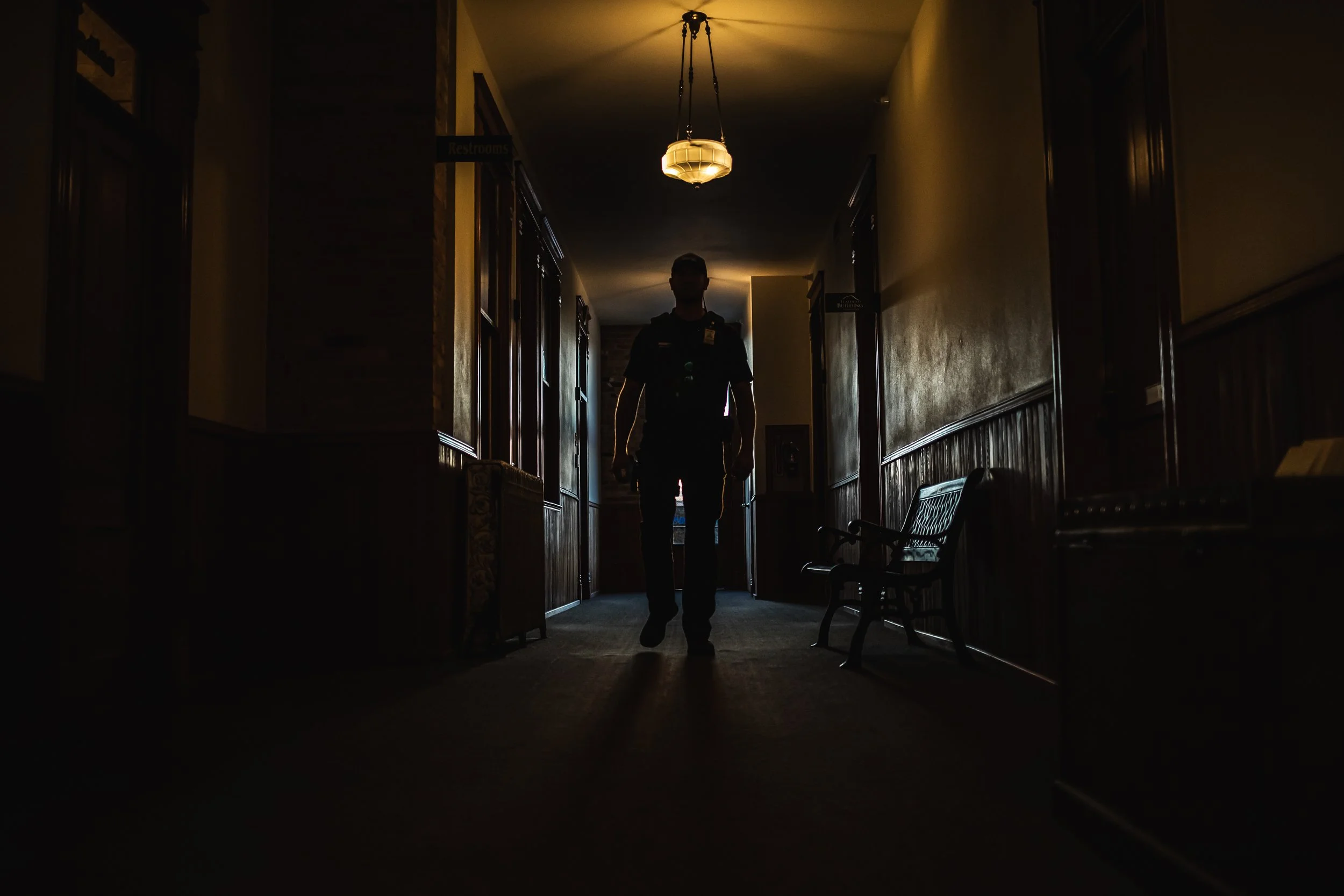 A dark hallway with a single person walking toward the camera, backlit by light at the end of the hall, with wooden paneling and a bench on the right side.