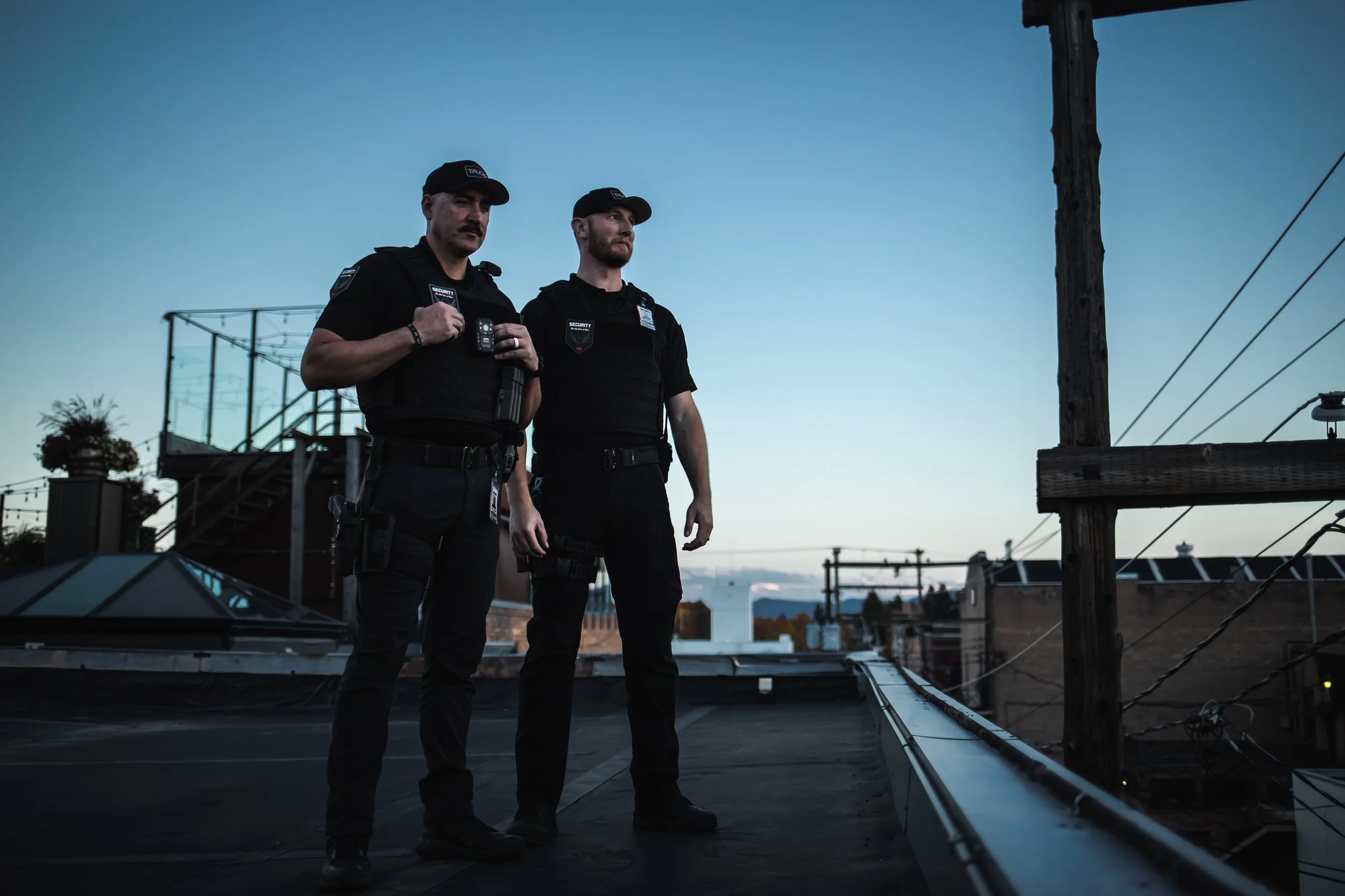 Two police officers standing on a rooftop at dusk, looking into the distance, with electrical wires and buildings in the background.