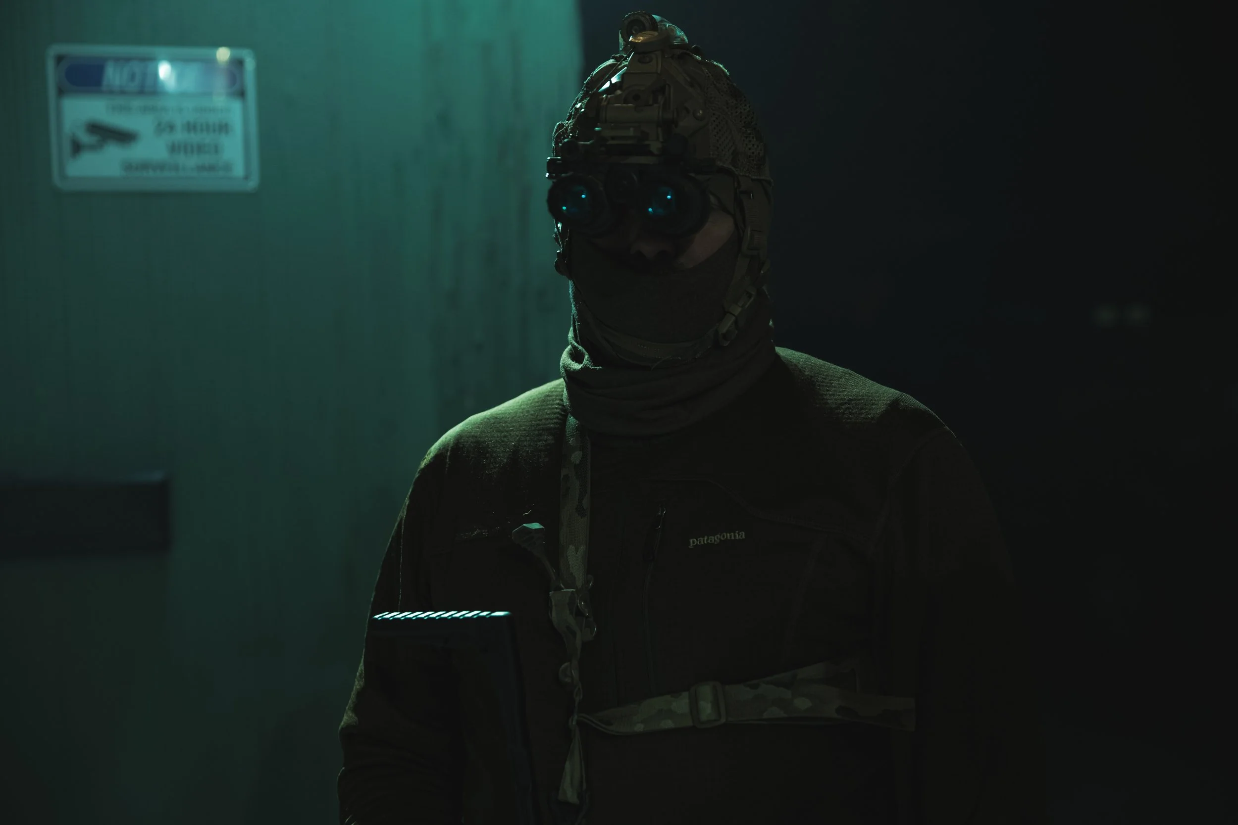 A person is wearing tactical gear, including a helmet with mounted night vision goggles, a face covering, and a dark: Patagonia jacket. The background is dim with a sign on the wall that says 'NOT' and appears to be a security or surveillance area.