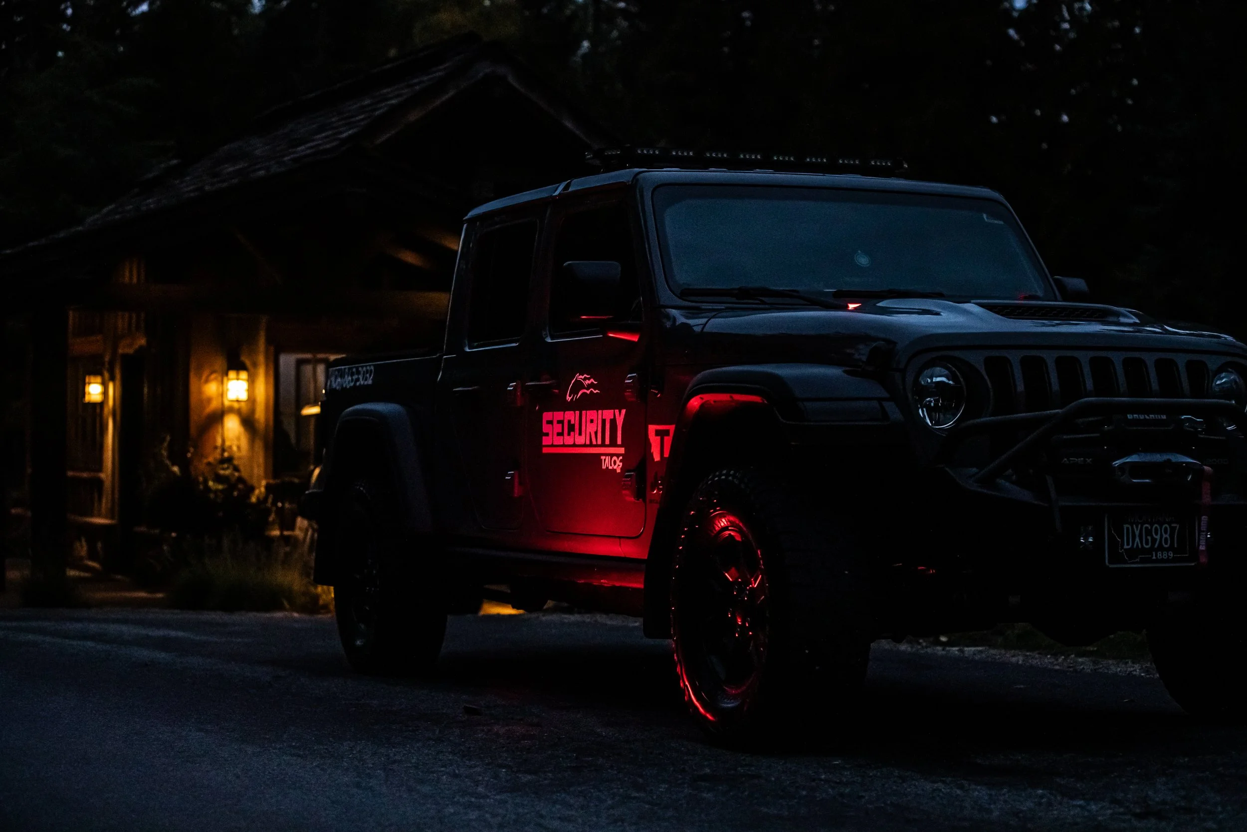 A black security patrol vehicle parked outside a wooden cabin at night, with illuminated red security decals on the side and dim cabin lighting in the background.