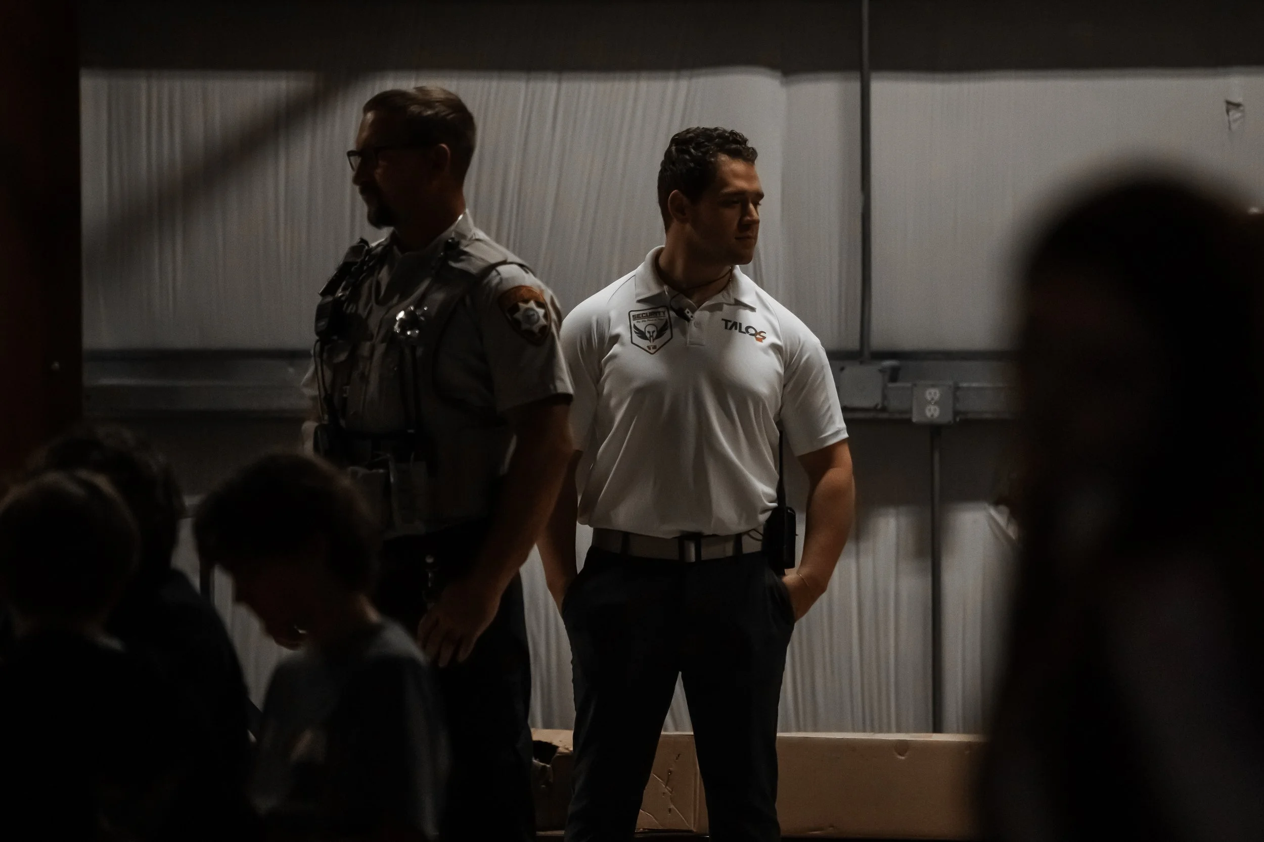 Two security officers standing in a dark room with children in the foreground, one in a sheriff's uniform and glasses, and the other in a white polo shirt with patches, looking off to the side.
