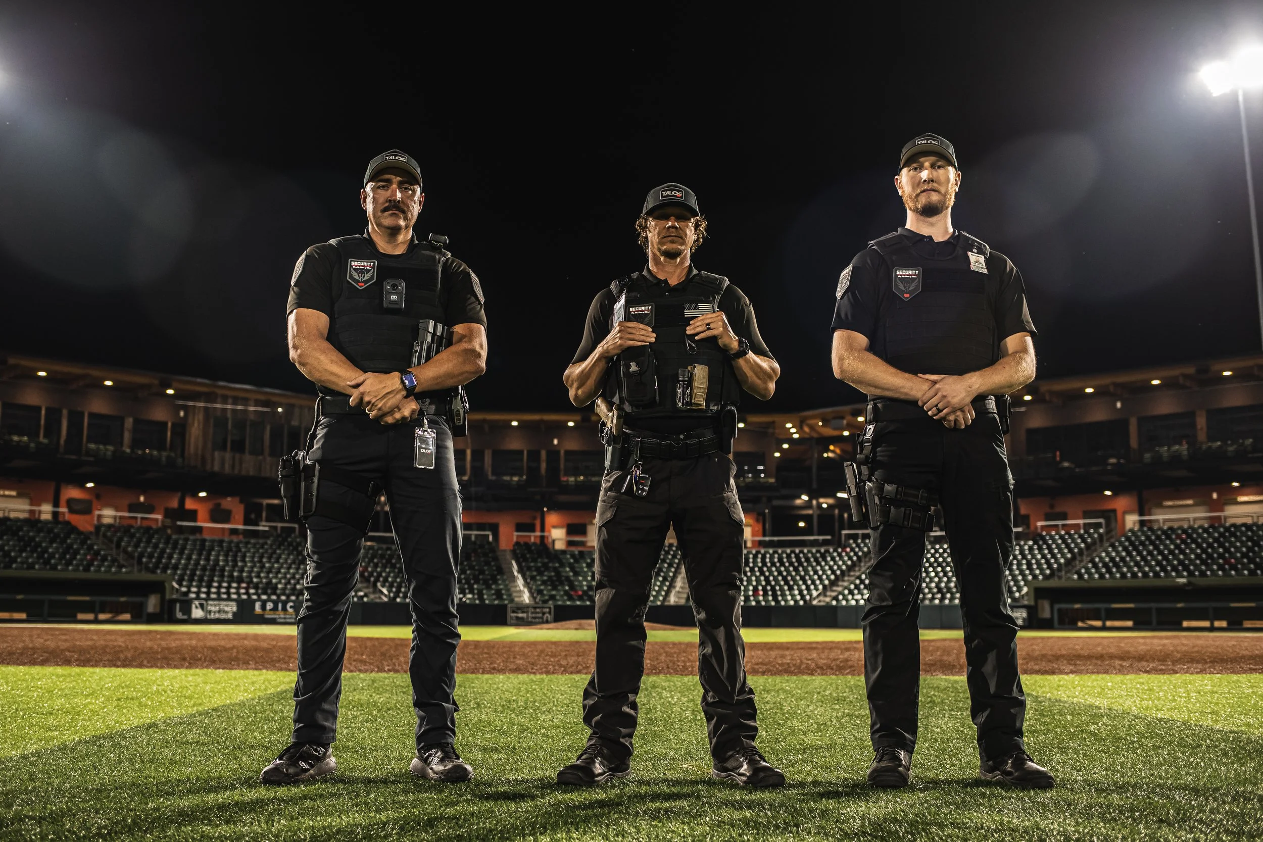 Three police officers in tactical gear standing on a baseball field at night.