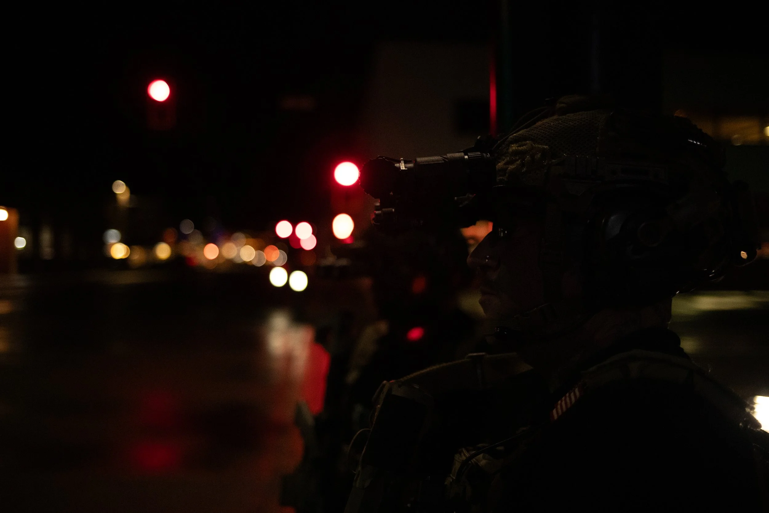 A silhouette of a soldier in uniform wearing a helmet with night-vision goggles, standing against a dark city street background with blurred red and white lights.