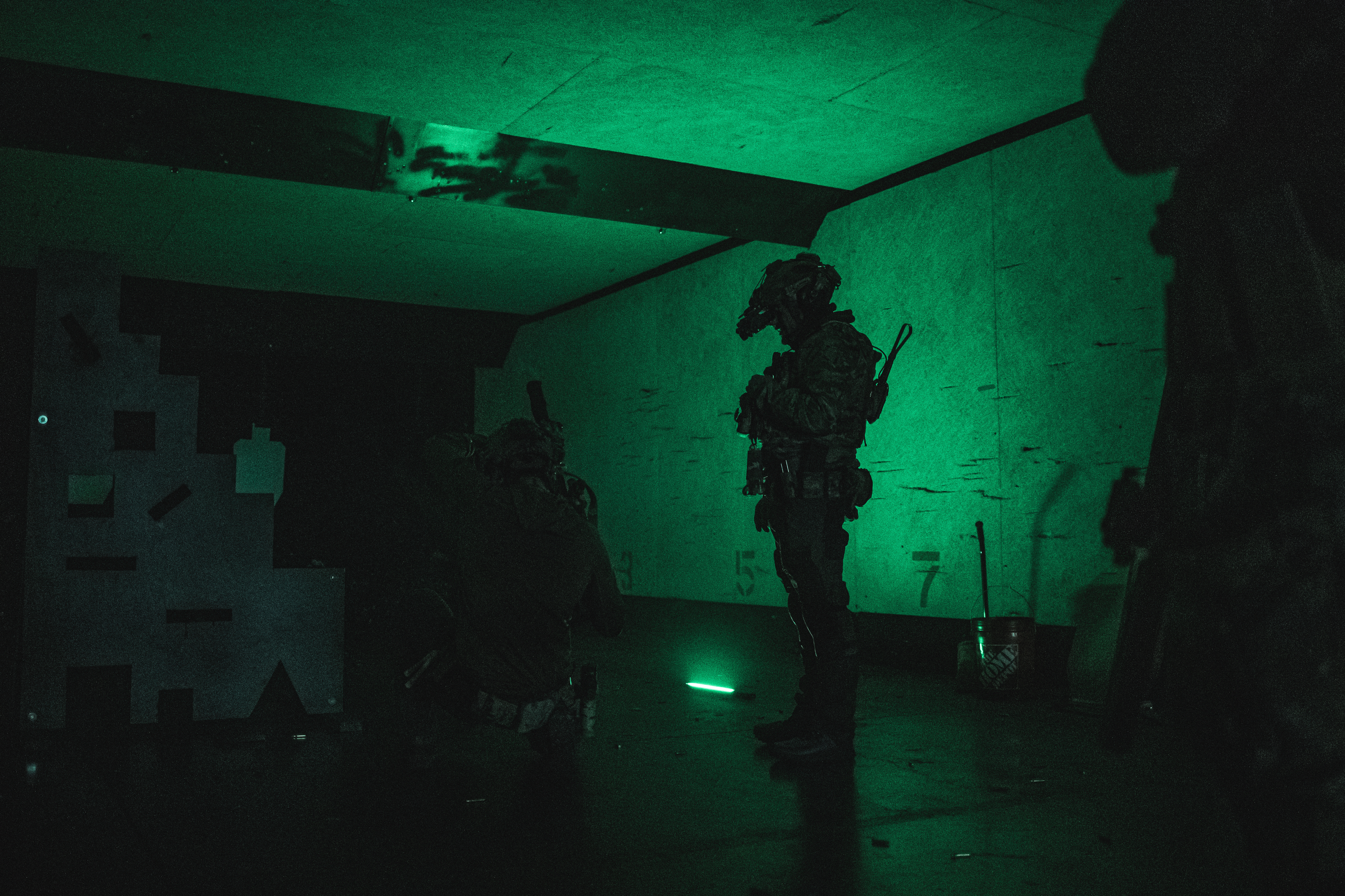 Three soldiers in tactical gear inside a dimly lit room with green lighting, one is standing while the others are kneeling, surrounded by various equipment and targets on the wall.