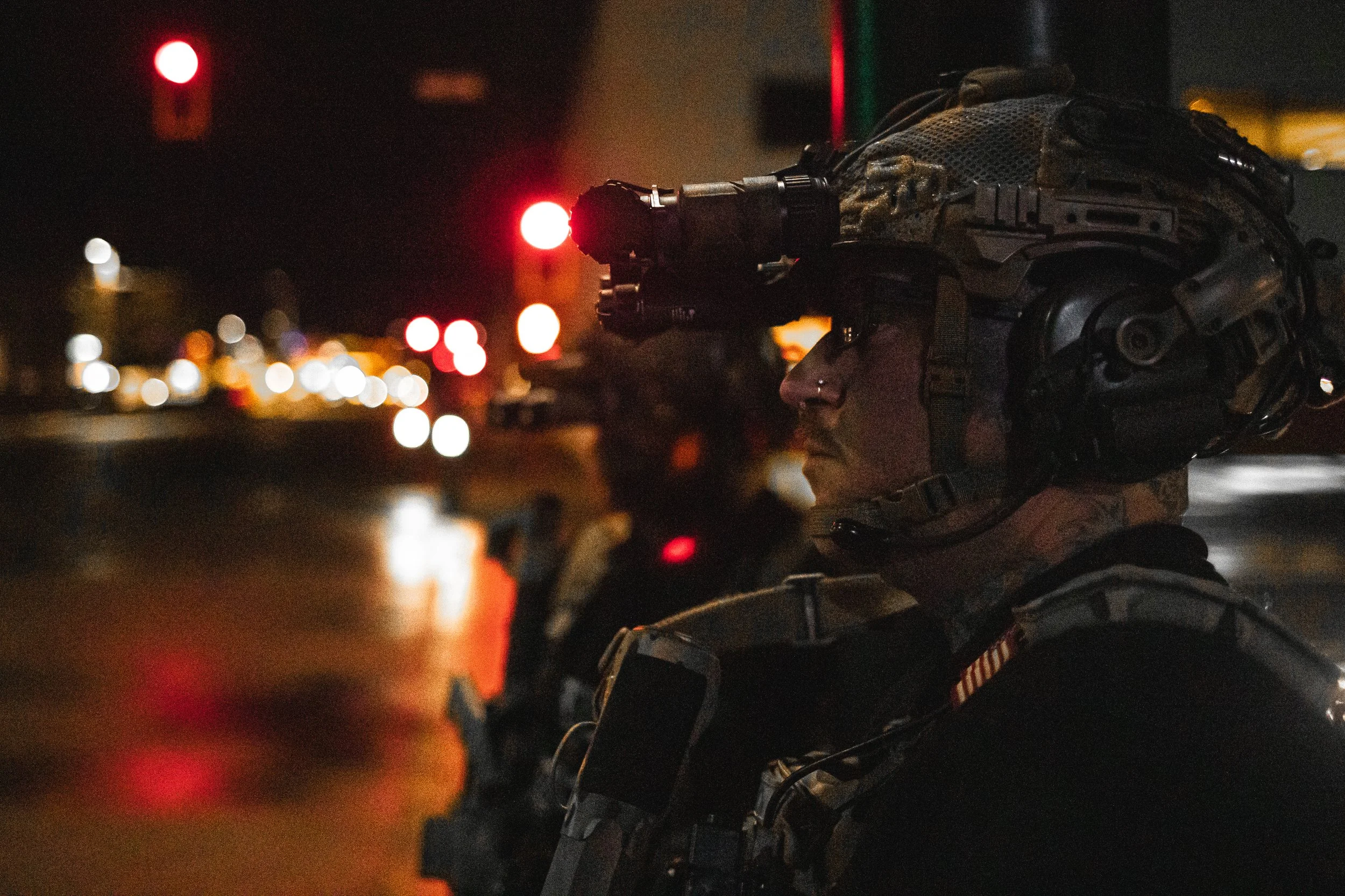Military person wearing tactical gear and helmet equipped with night vision goggles and a flashlight, standing in a city at night with blurred city lights in the background.