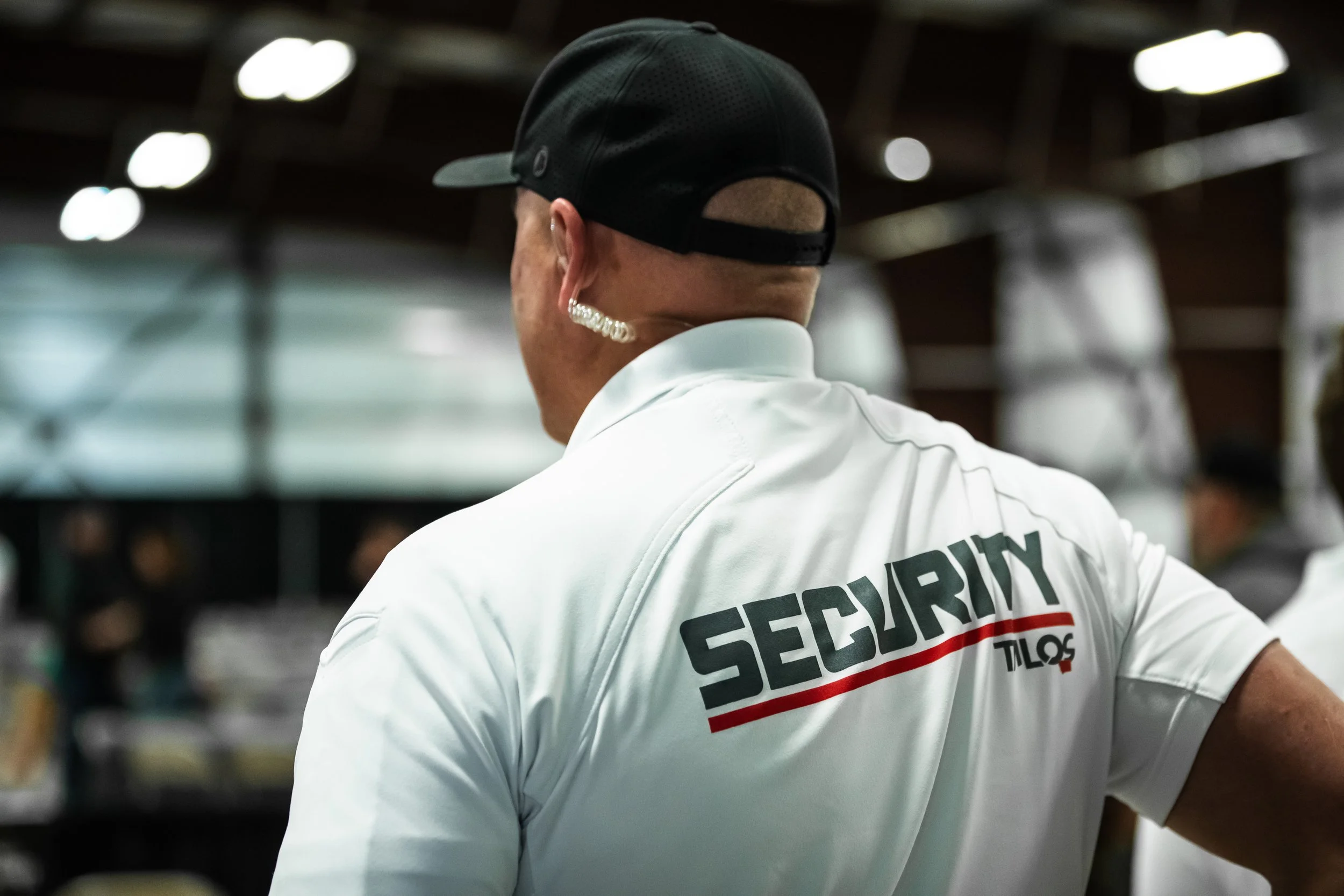 Security personnel wearing a white shirt with 'SECURITY' printed on the back, black cap, earring, and diamond earrings, standing in an indoor setting with blurred background.