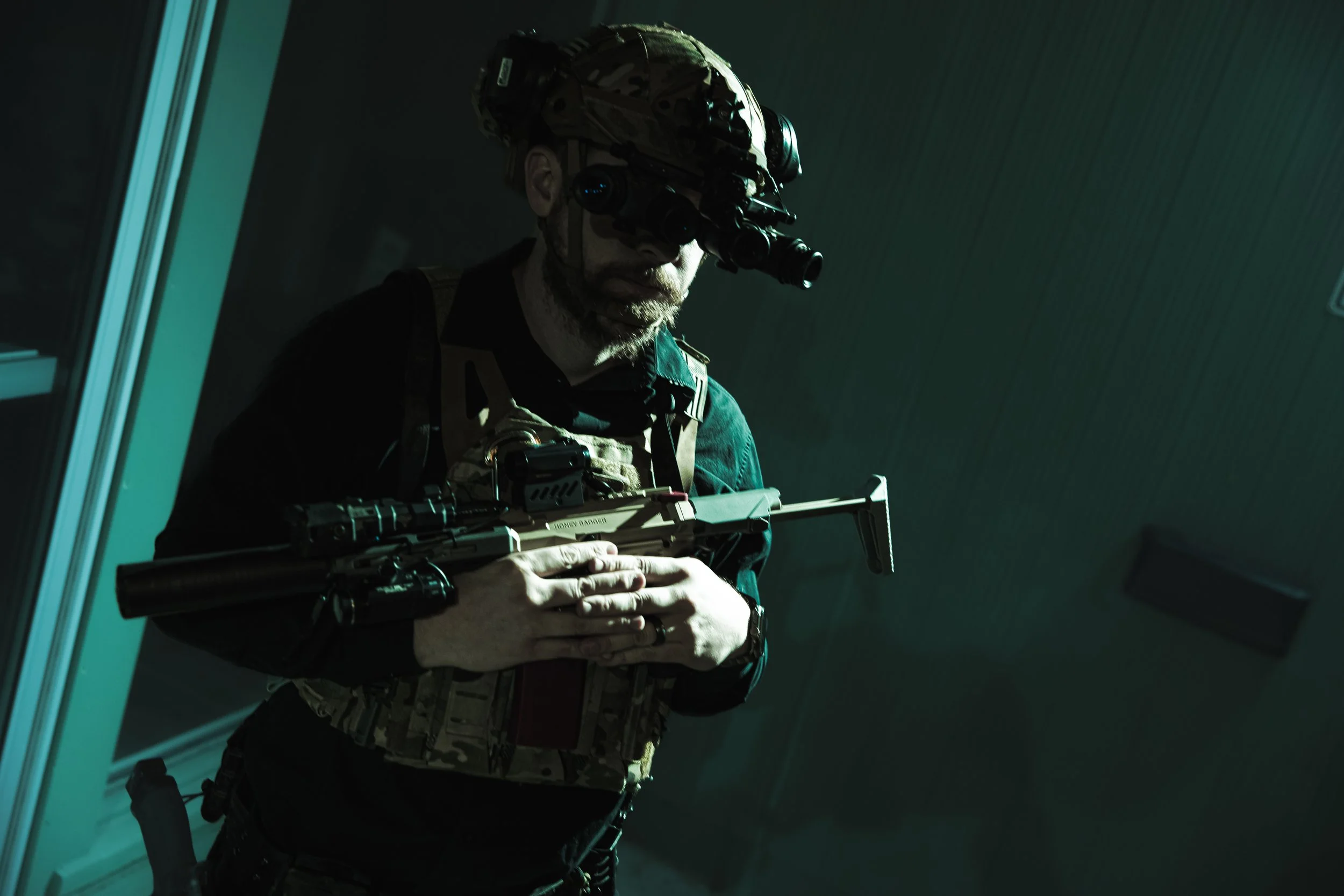A soldier or special forces operative wearing tactical gear and a helmet with night vision goggles, holding a sniper rifle, indoors in a dark setting.