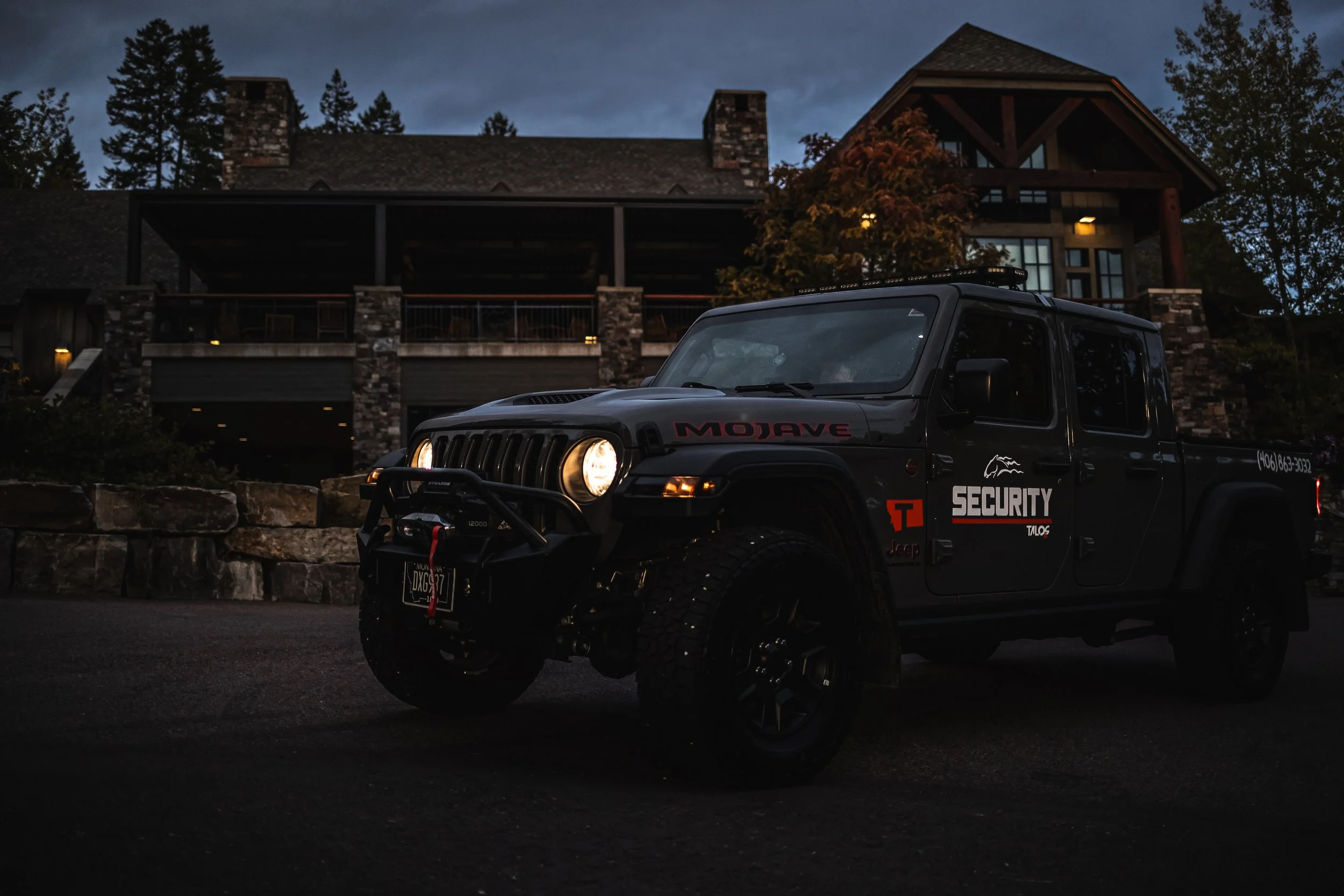 A black Jeep security vehicle with its headlights on parked in front of a large house at dusk or night.