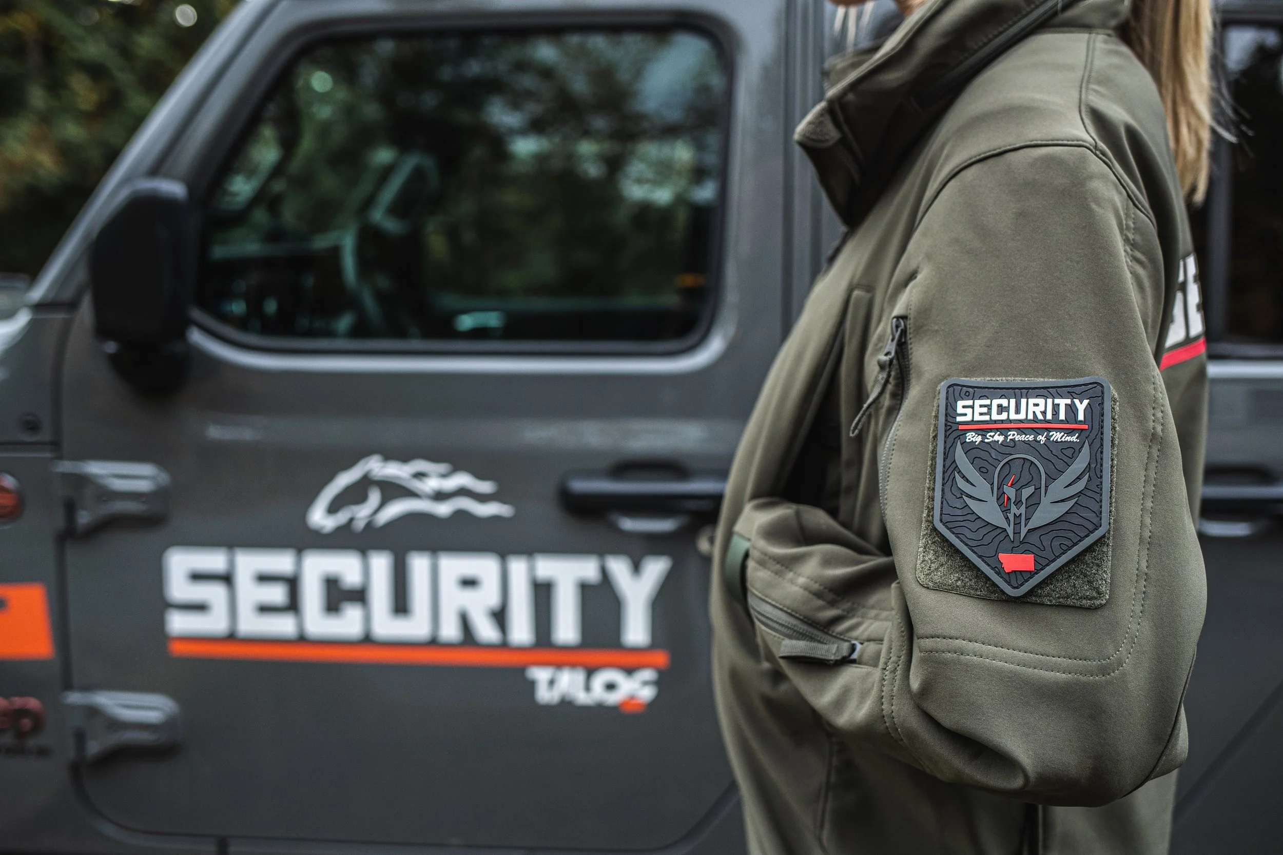 A security guard in a green jacket with a security patch standing beside a security vehicle with the label 'SECURITY Talos' and a wolf logo.