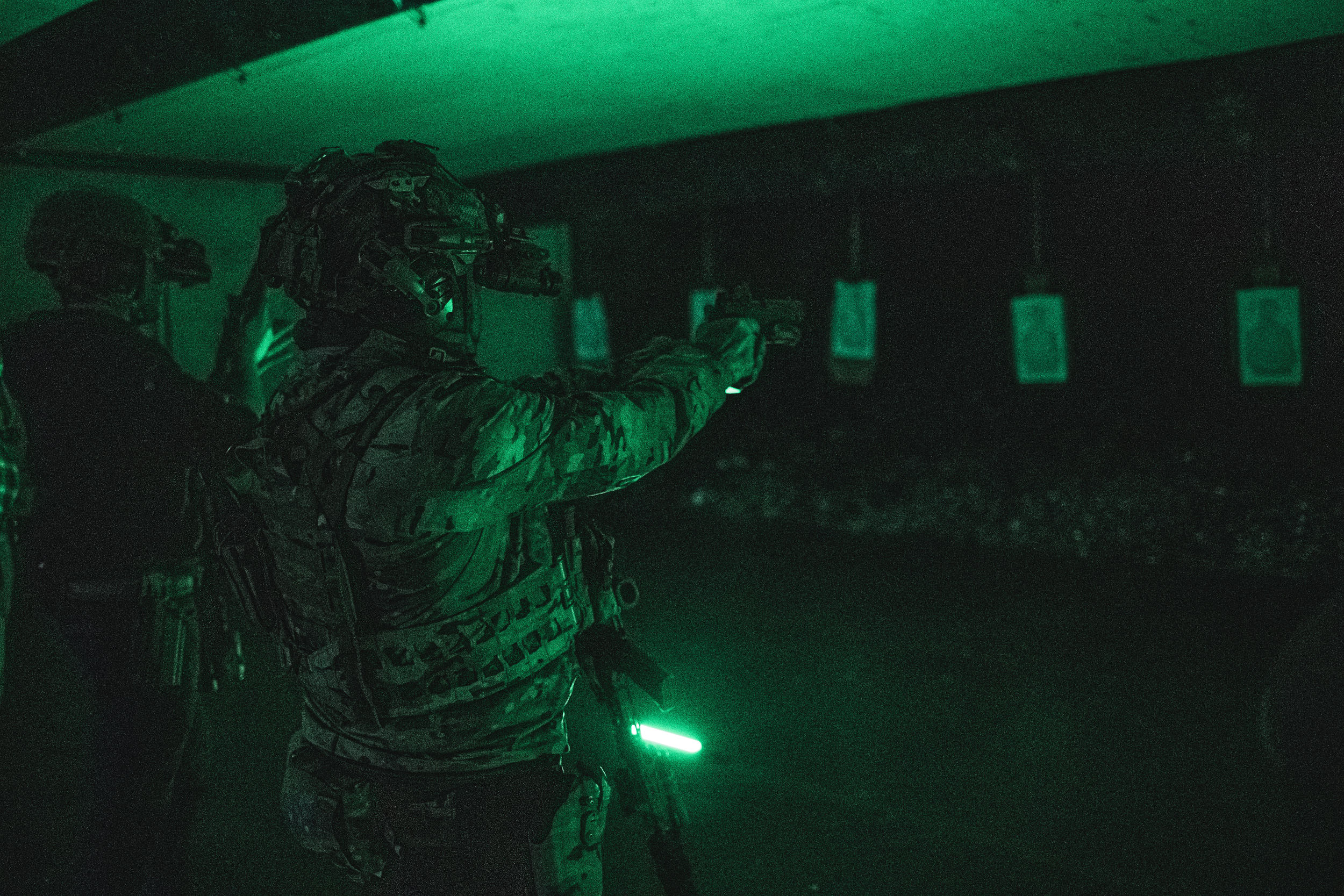 Military personnel in tactical gear aiming at targets in a dark shooting range with green lighting.