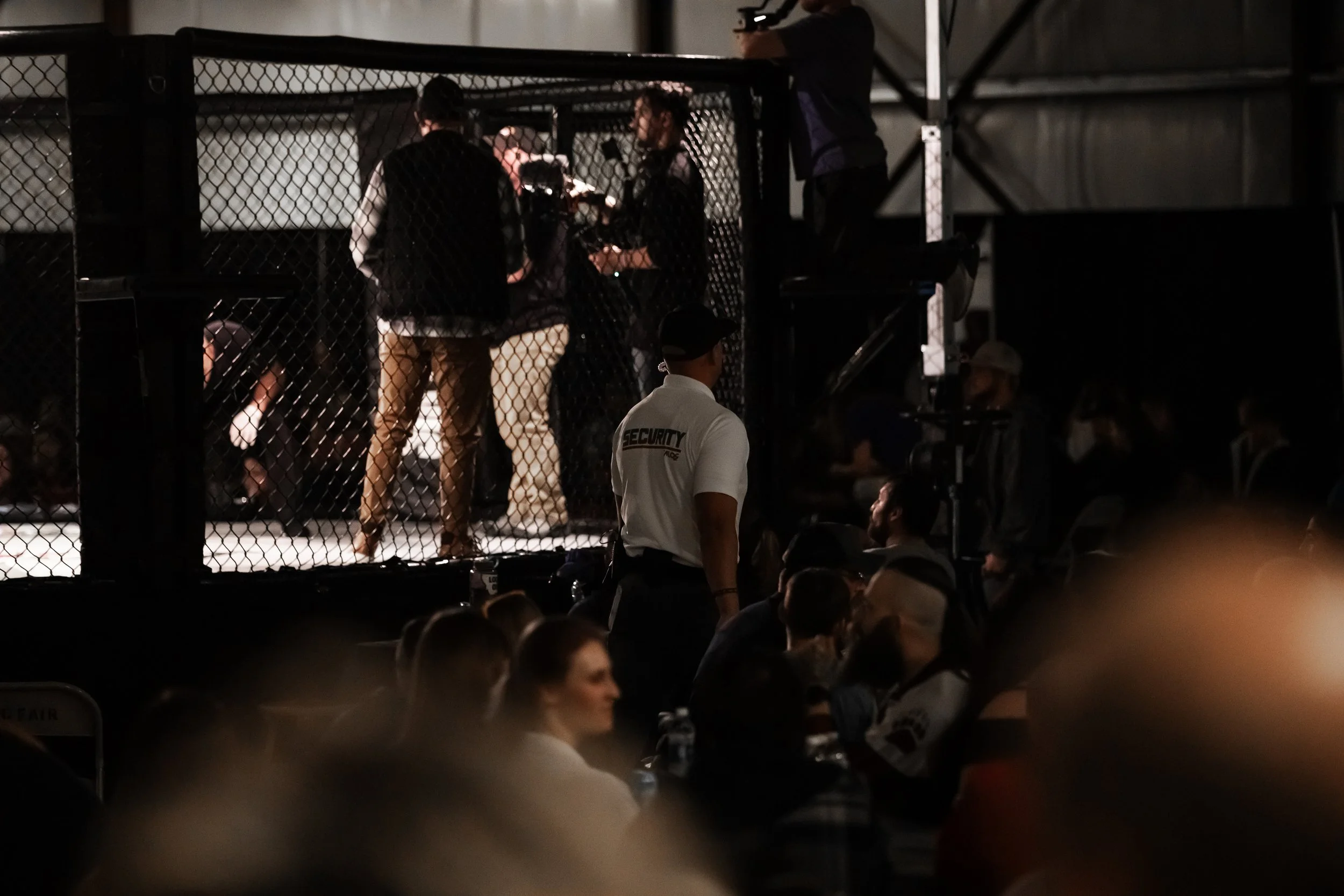 Mixed martial arts event inside a cage, with fighters, trainers, and officials, viewed from the audience with security personnel visible.