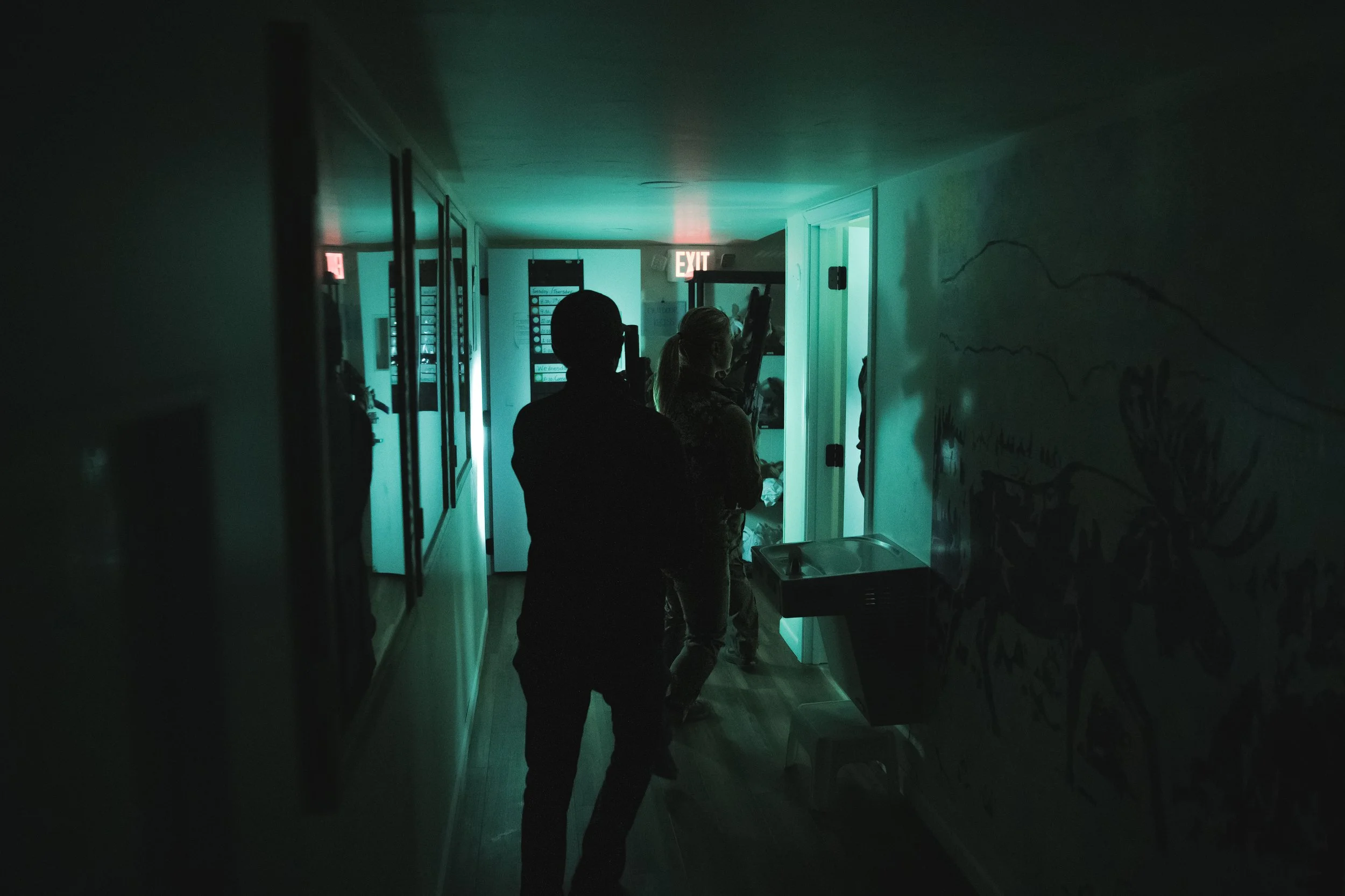 Silhouettes of two people walking in a dimly lit corridor with teal lighting, an exit sign overhead, and a mural on the wall to the right.