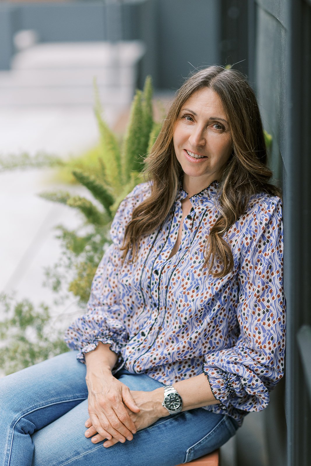 Woman with long brown hair wearing a colorful floral blouse and jeans sitting outdoors next to a dark wall and green plants.
