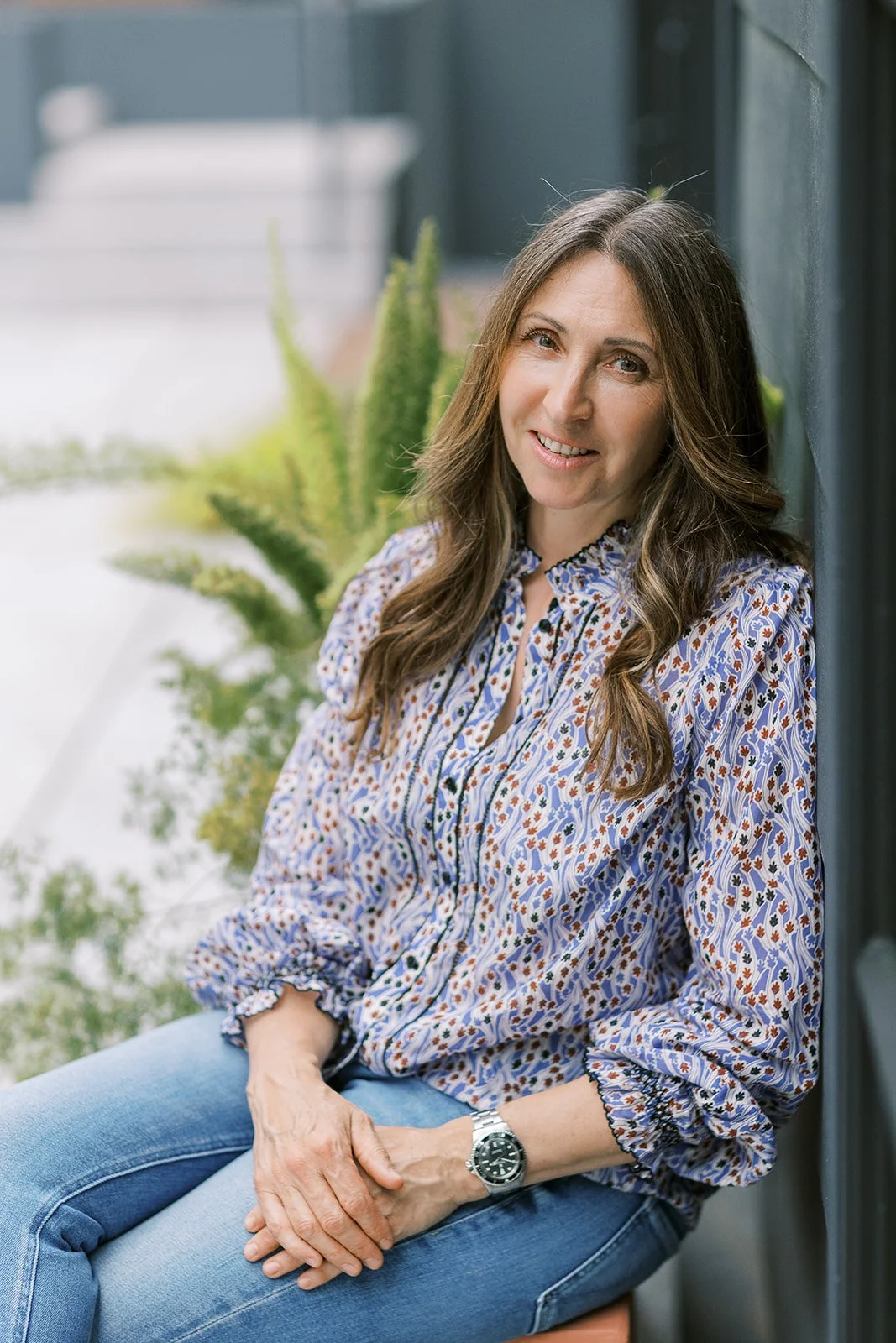 A woman with long brown hair wearing a colorful patterned blouse and jeans, sitting against a dark wall with an outdoor setting and green plants behind her, smiling at the camera.