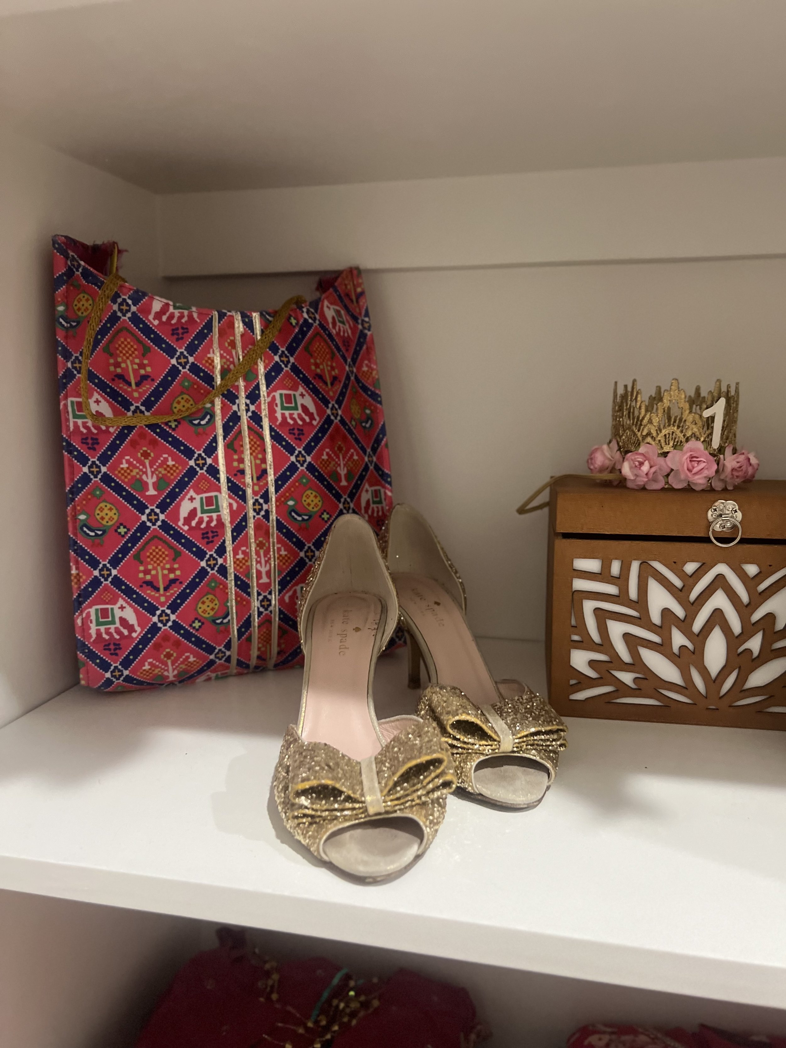 Pair of gold glitter high heels with bows, a colorful patterned gift bag, and a small wooden box with pink roses and a gold crown on top.
