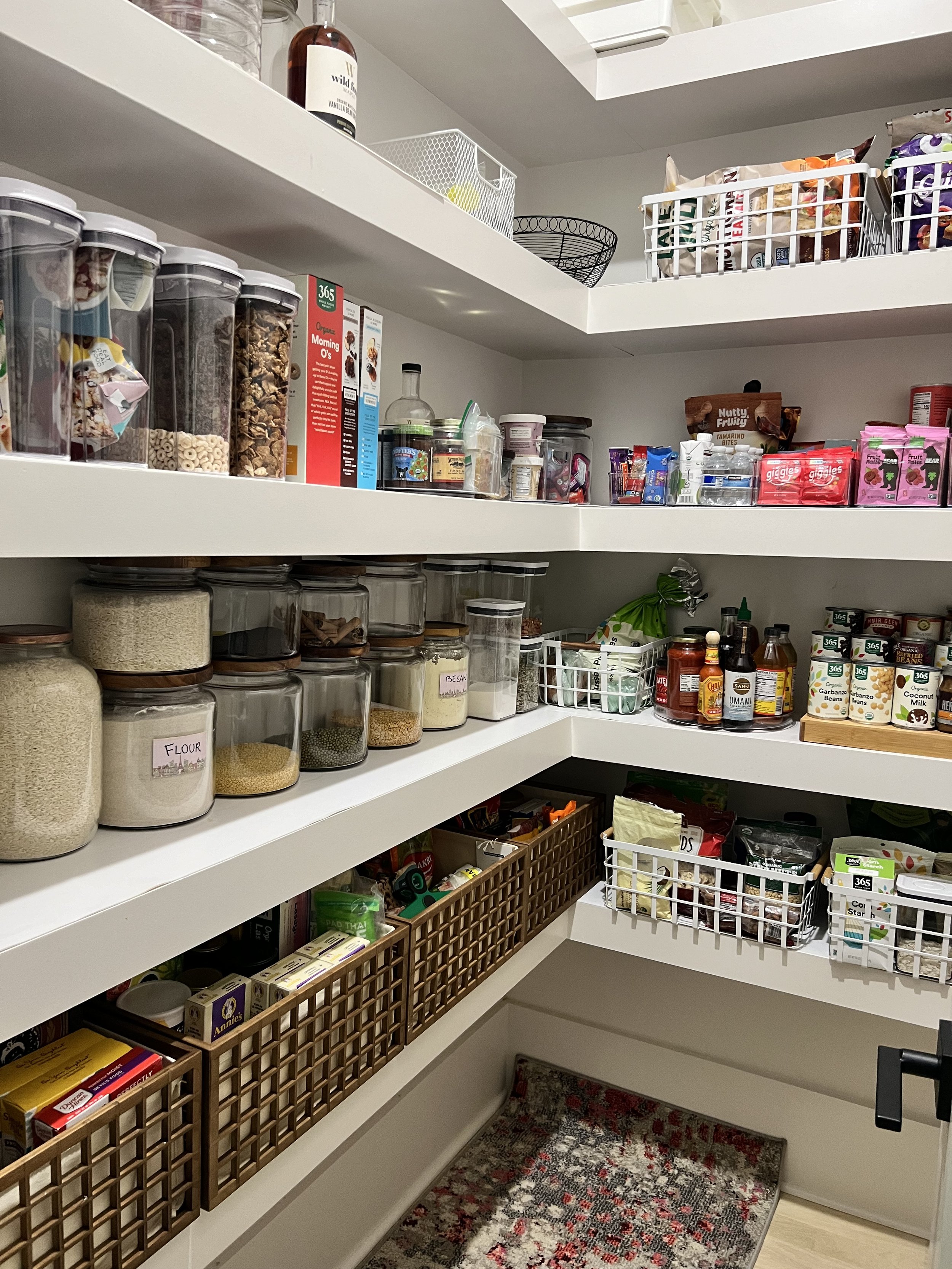 A well-organized pantry with shelves holding various food items, jars, bottles, and boxes, including grains, snacks, condiments, and baking ingredients.