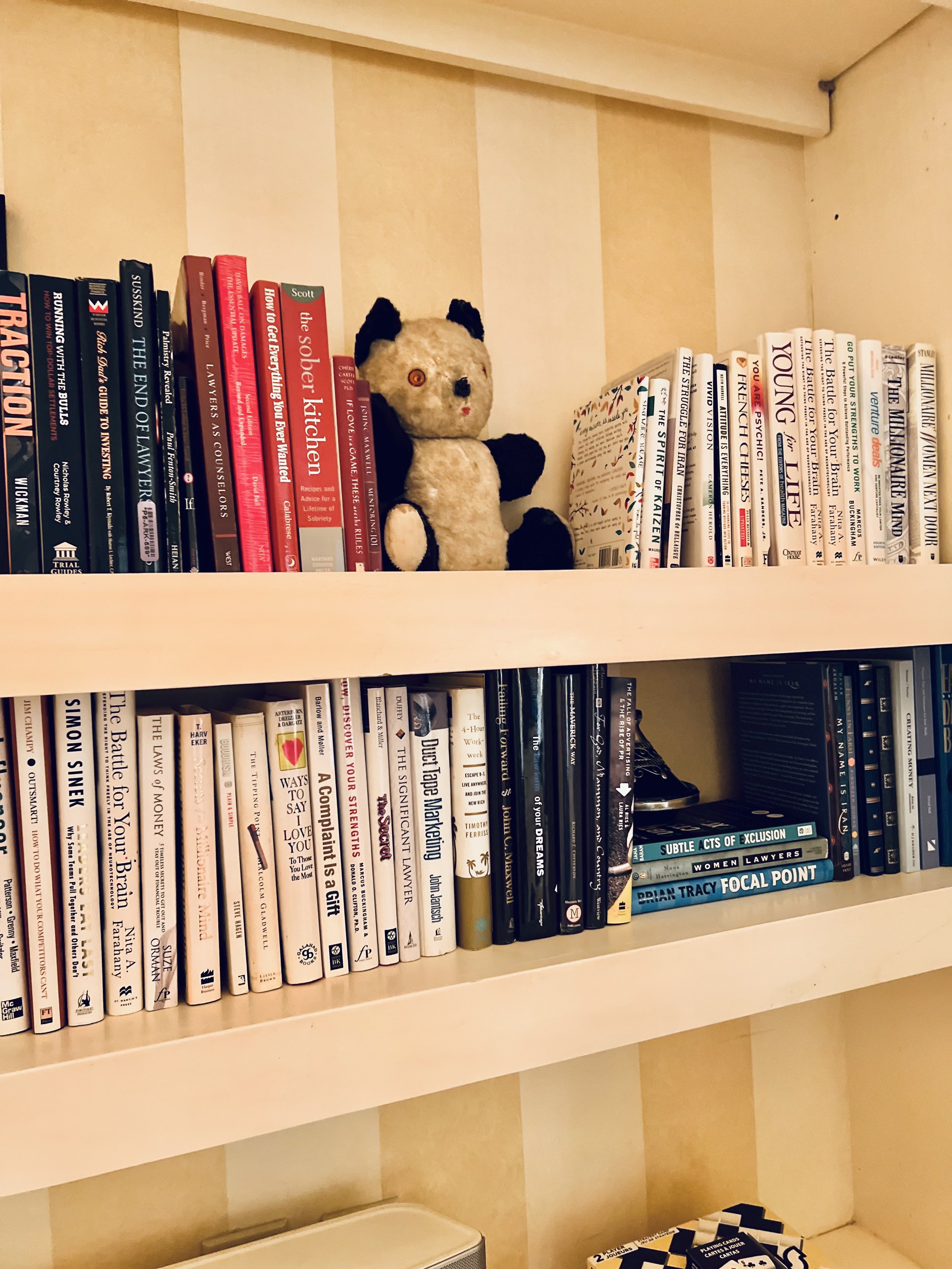 A beige bookshelf with two shelves holding books and a plush panda toy. The top shelf has red and black books and a panda plush toy, while the middle shelf contains mainly white and black books, including three stacked ones.