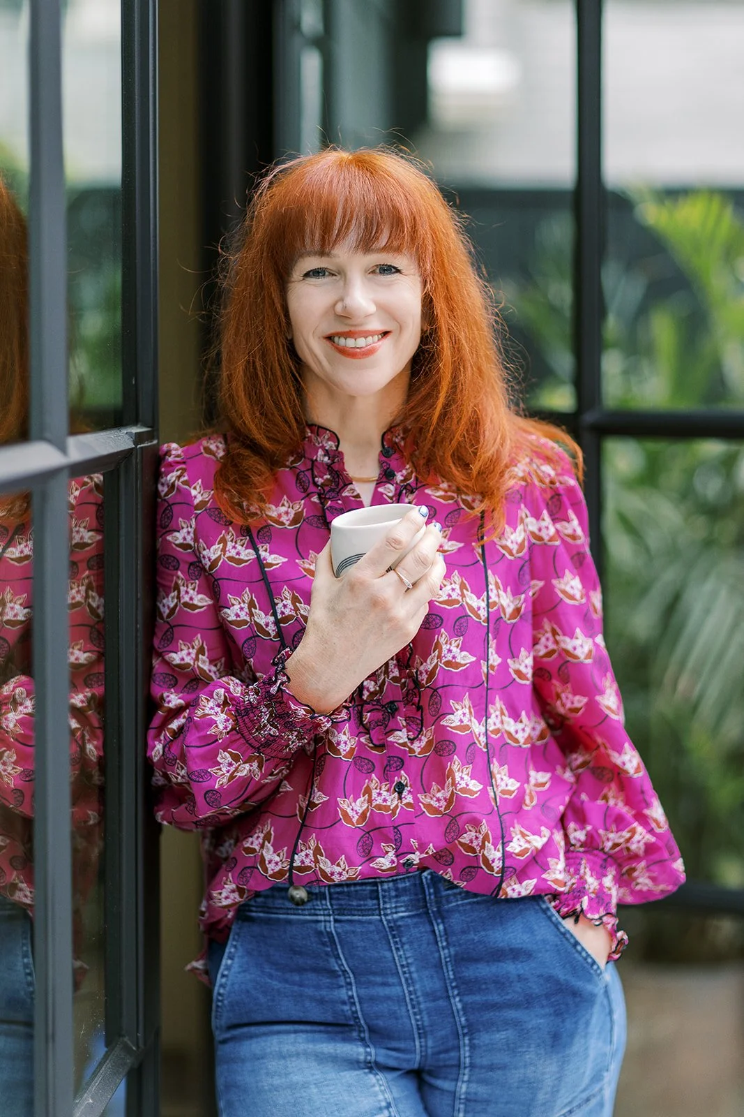 A woman with red hair wearing a pink floral blouse and blue jeans, holding a white mug, standing near a glass window with greenery in the background, smiling.