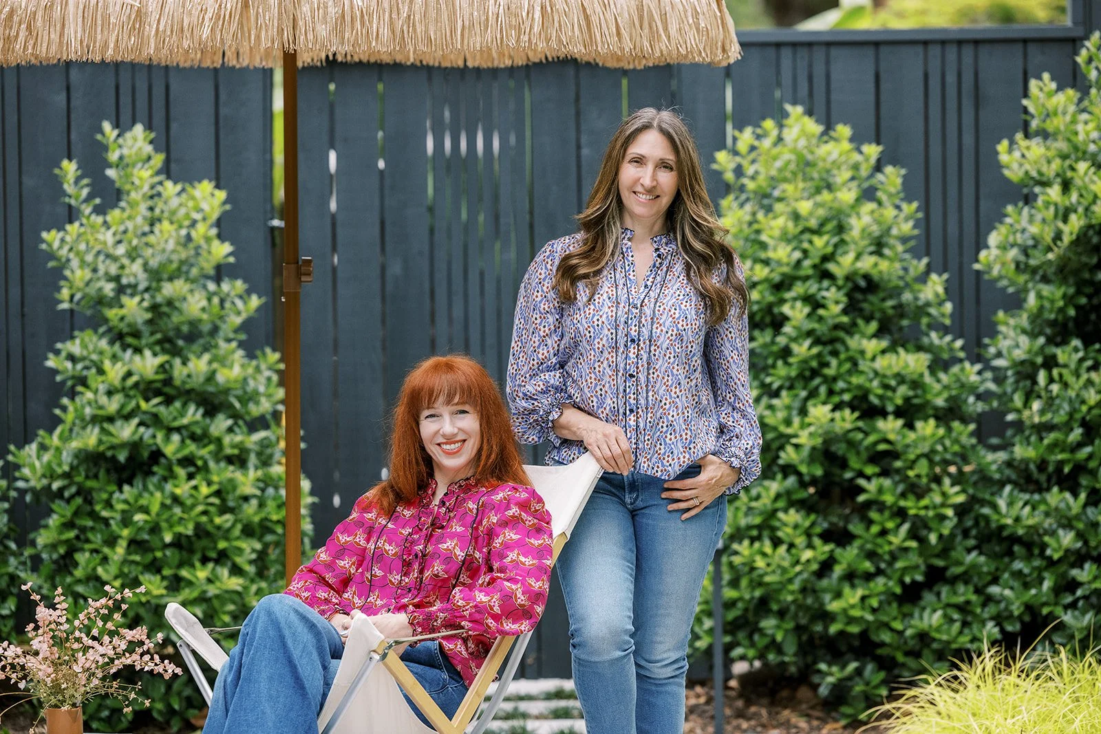Two women in a garden under a thatched umbrella, one sitting in a chair with red hair and the other standing with brown hair, surrounded by greenery.
