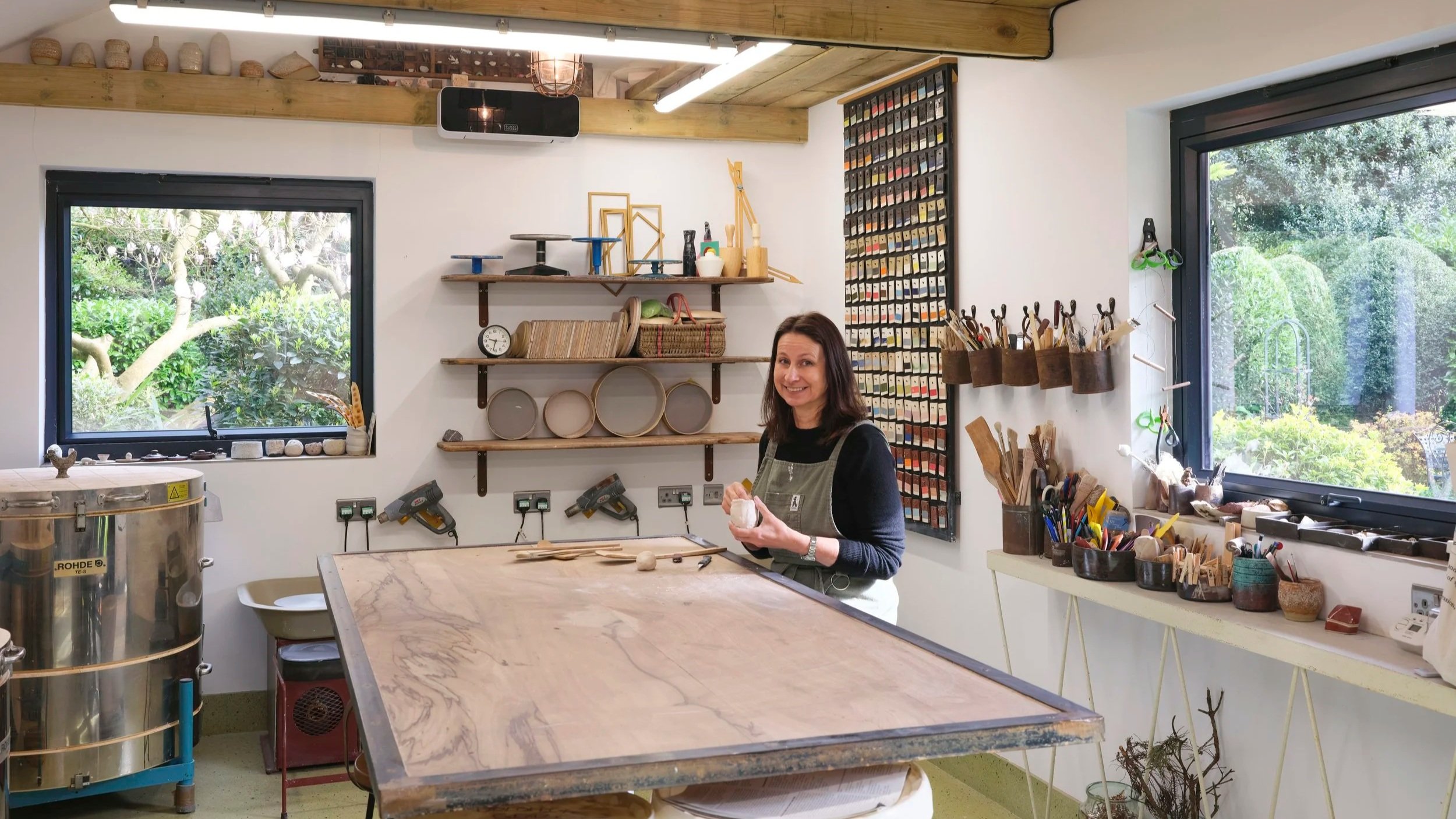 Marija Bichan working in The Ugly Barn pottery workshop surrounded by kilns, handmade ceramics, colour swatches, and pottery tools, with views of the garden outside in Ditchling, Sussex.