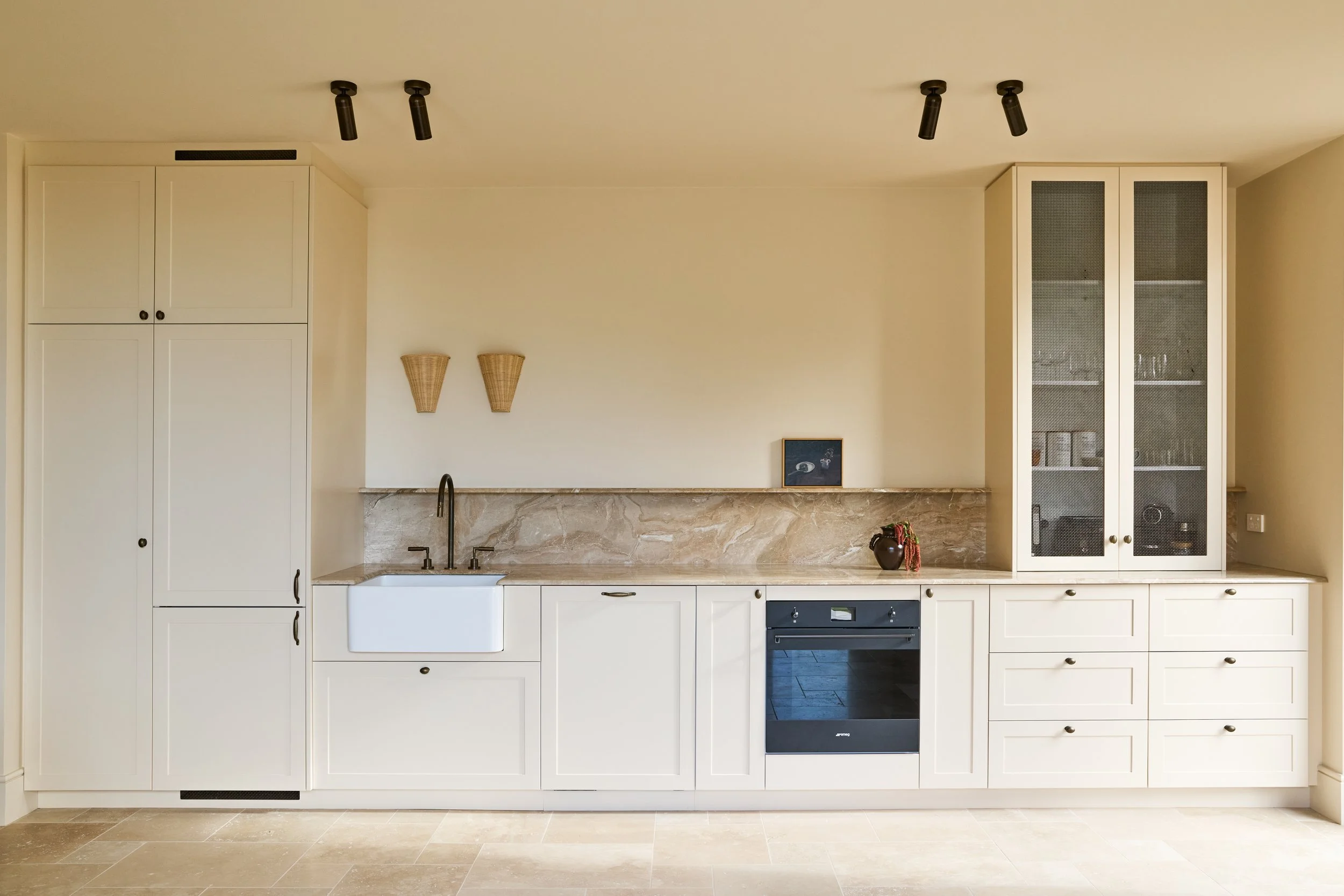 Modern kitchen with white cabinets, black oven, marble countertop, and sink, decorated with a black vase and a framed picture on the counter, with two woven wall sconces and black ceiling spotlights.