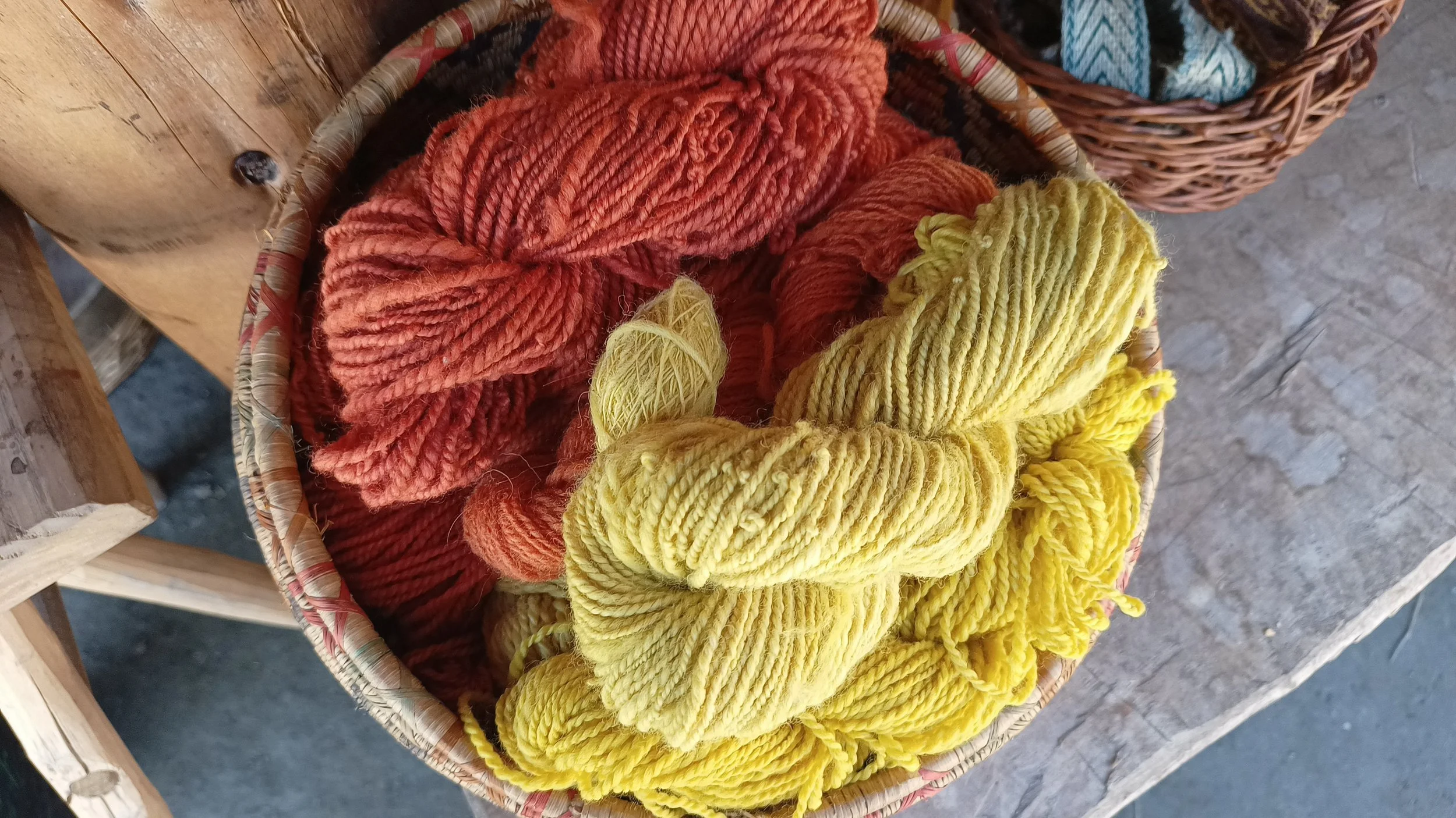A woven basket filled with skeins of wool yarn in warm shades of rusty orange, golden yellow and pale yellow, displayed on a wooden surface.