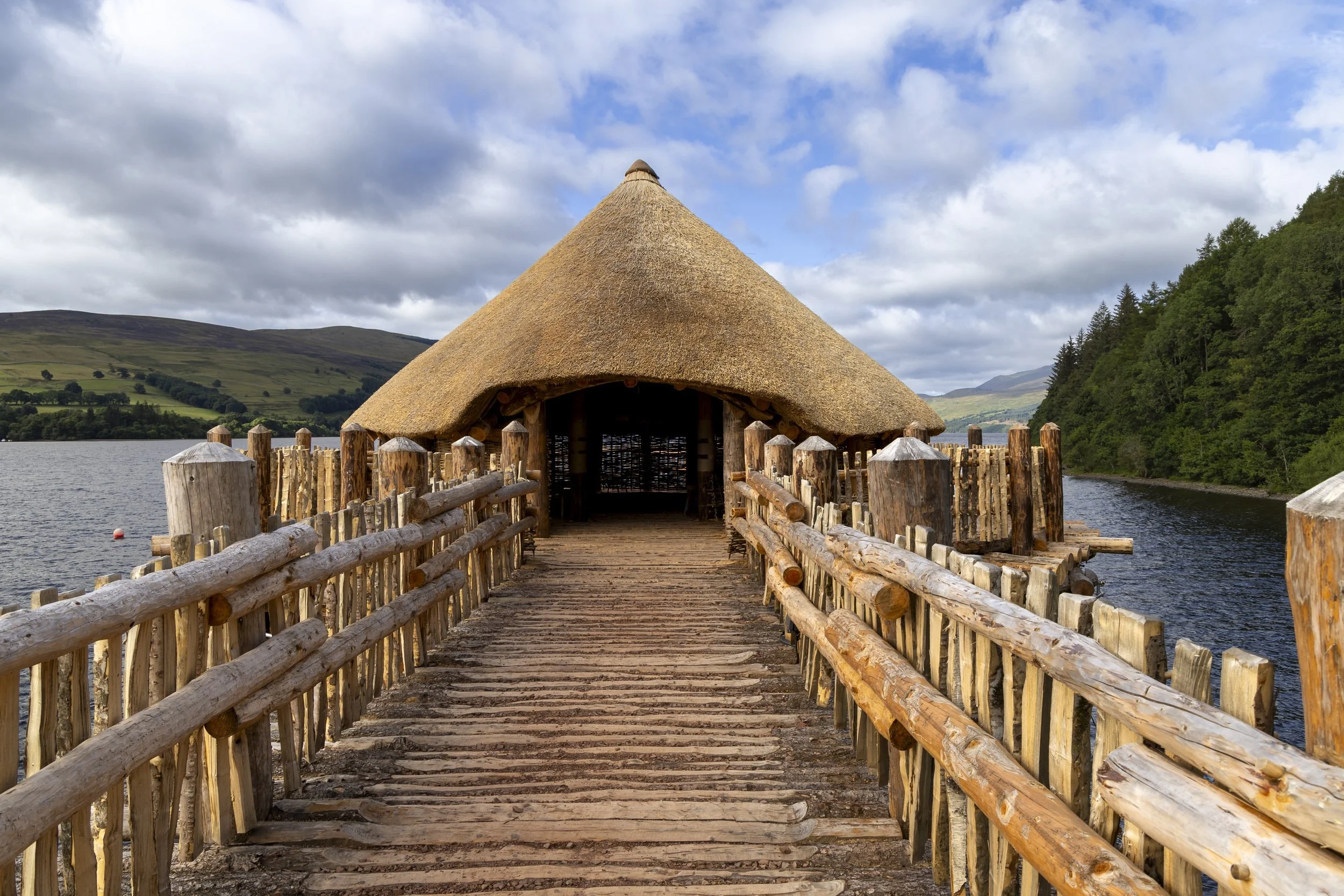 A reconstructed Iron Age crannog with a large conical thatched roof, reached by a wooden walkway over a Scottish loch, with wooded hills and mountains in the background.