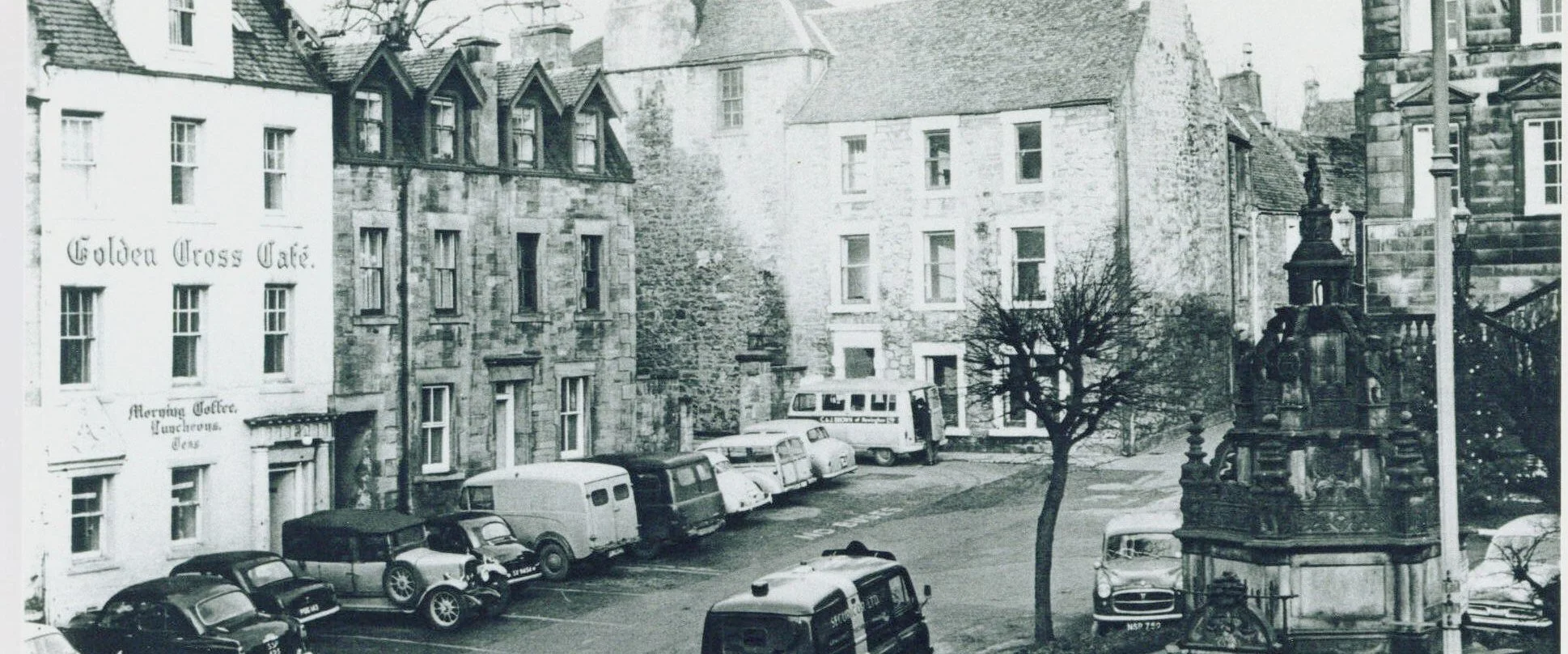 Historic photograph of Linlithgow Cross, showing buildings, parked cars and the town centre as it appeared in the past.
