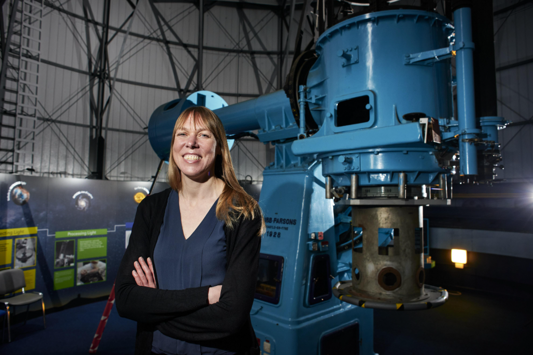 Professor Catherine Heymans, Astronomer Royal for Scotland, standing in front of a large blue telescope at an observatory.