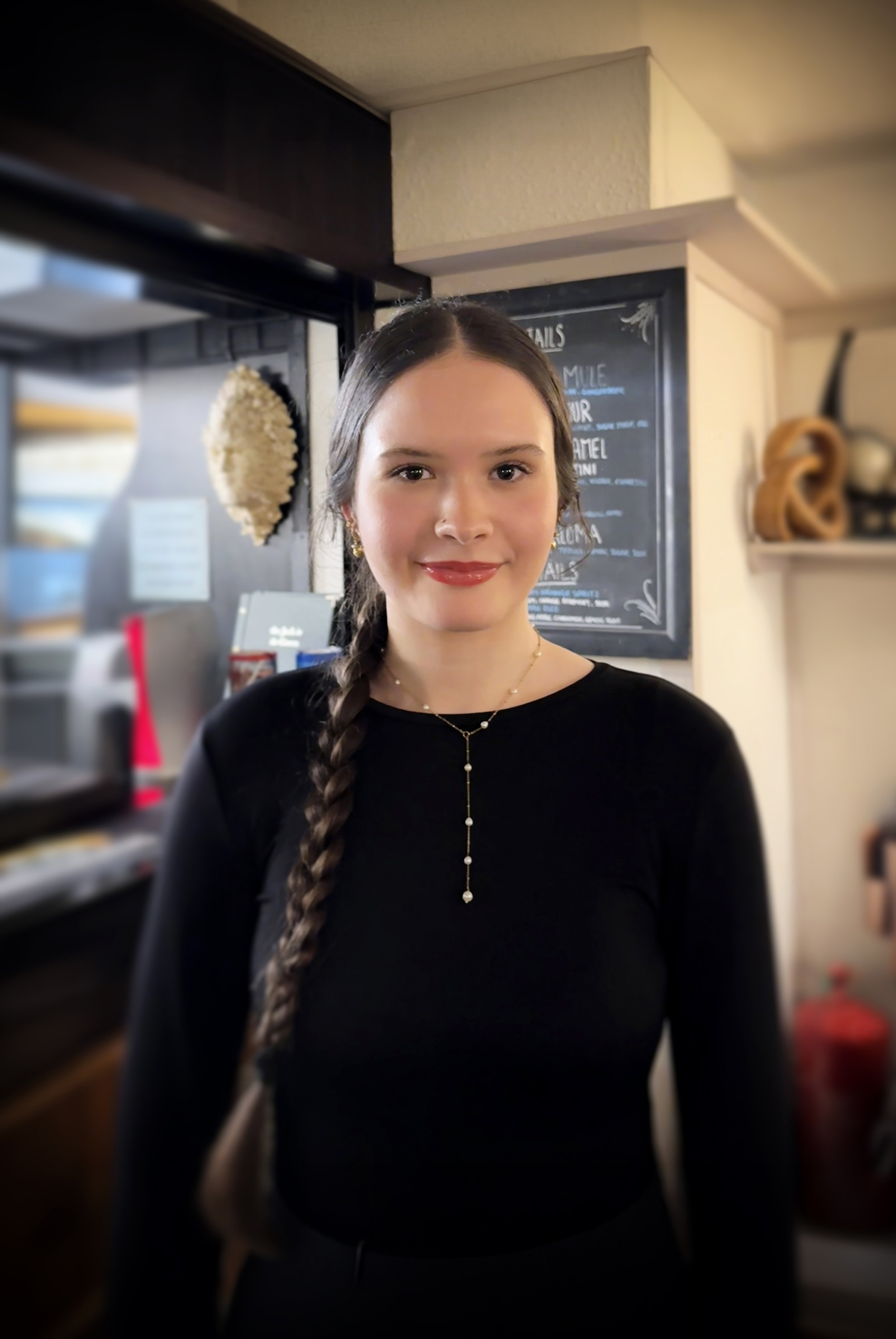 A woman with long red hair braided over her shoulder, wearing a black blouse and gold jewelry, standing in a dimly lit restaurant or bar.