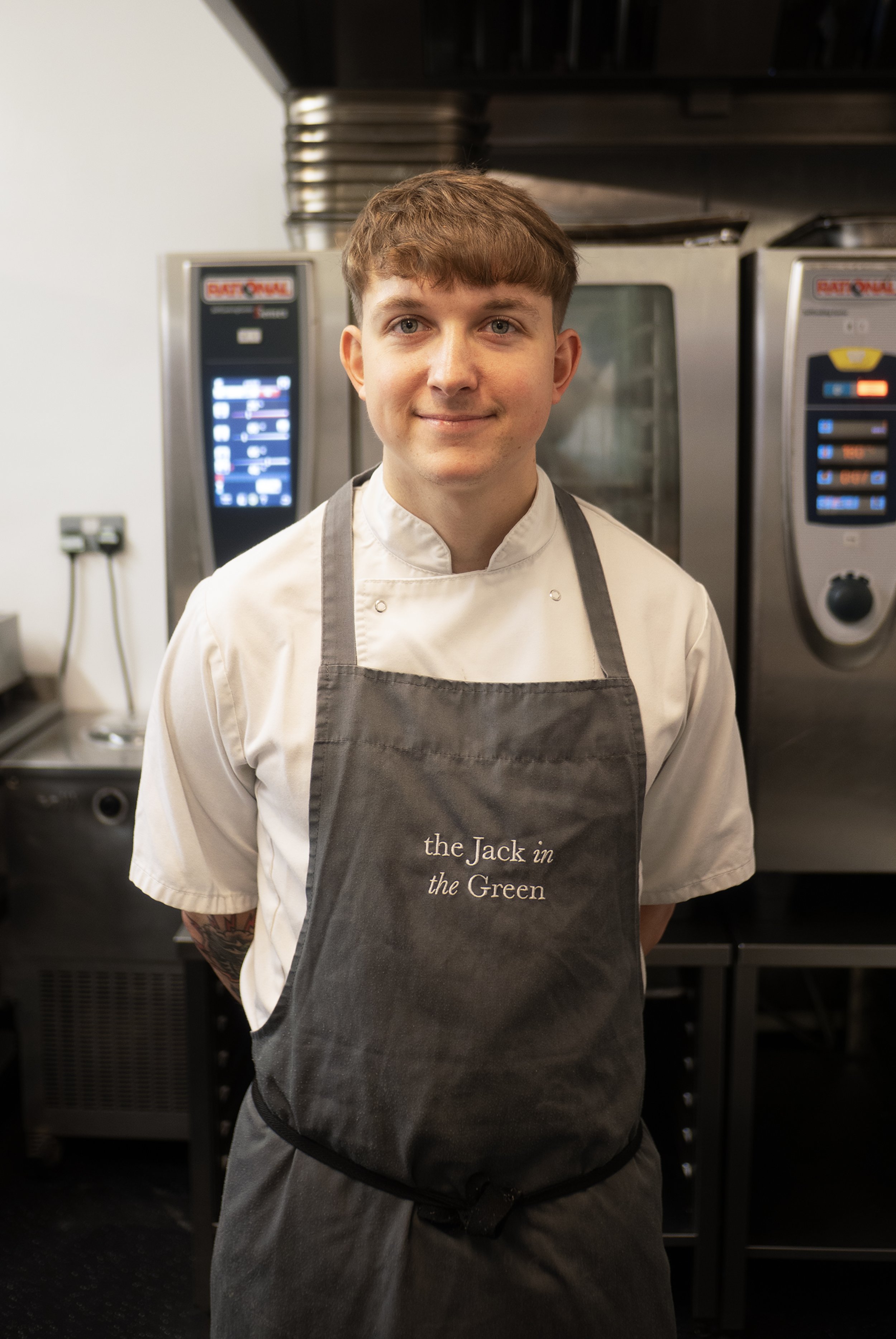 A young male chef wearing a white chef coat and a dark apron, smiling in a commercial kitchen with professional ovens in the background.