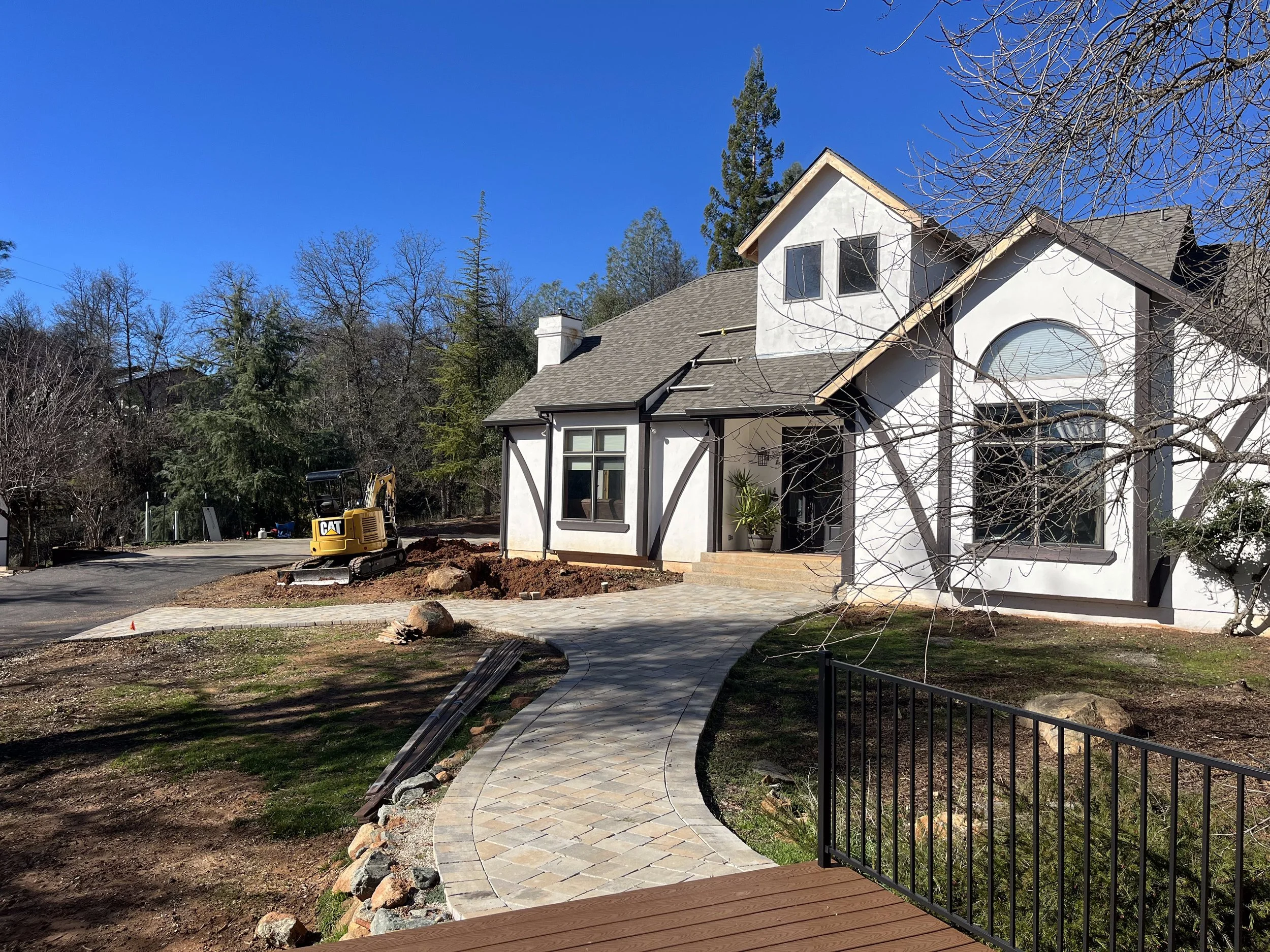 A house under construction with a curved stone walkway in front and a small excavator nearby. The house is white with dark trim and large windows, with some trees around it and a blue sky in the background.