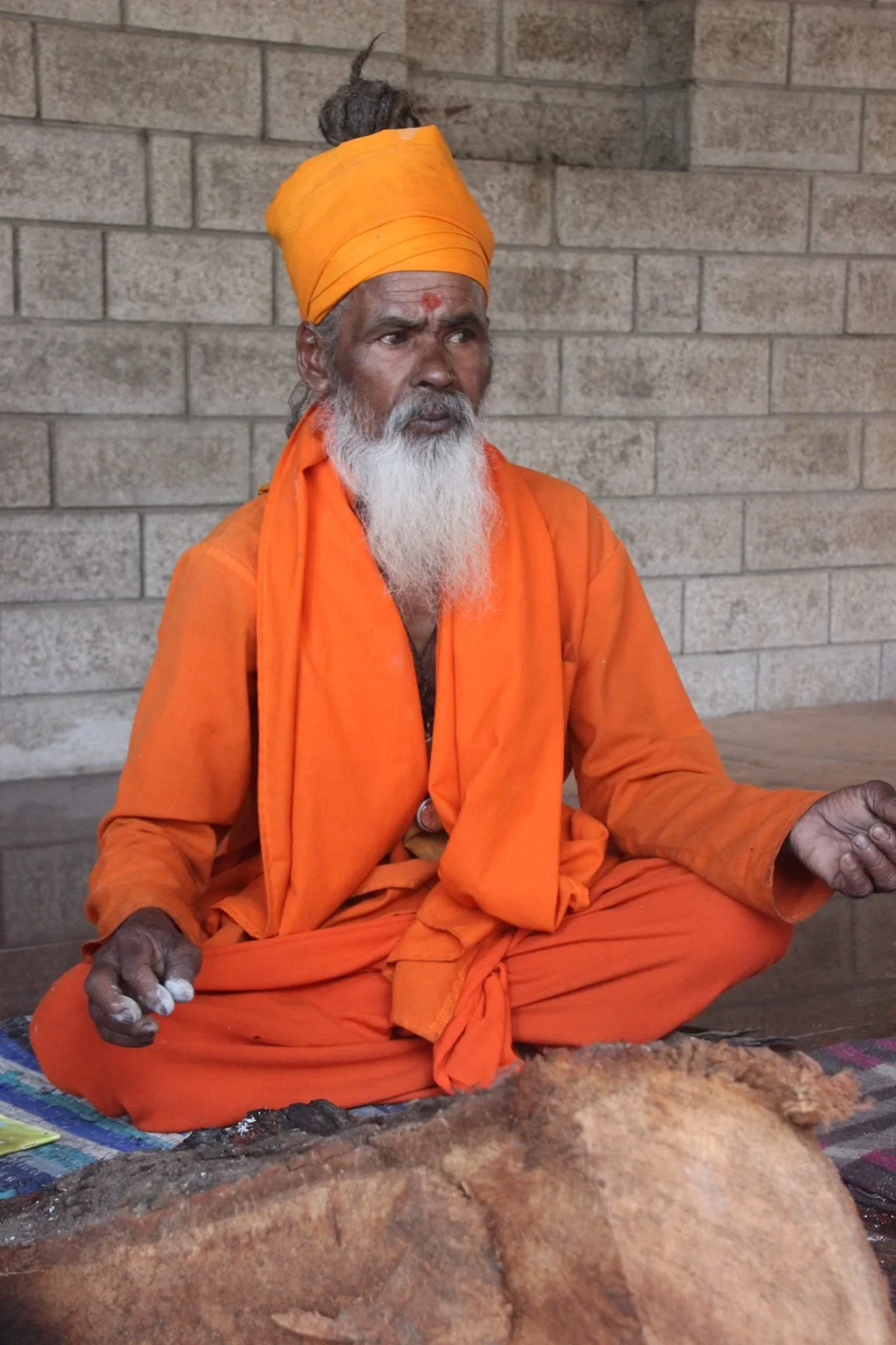 Ein älterer Mann mit grauem Bart und dunkler Haut sitzt im Lotussitz und trägt eine orange Robe und einen Turban. Er sitzt vor einer Steinmauer, möglicherweise in einem Tempel oder einer spirituellen Stätte.