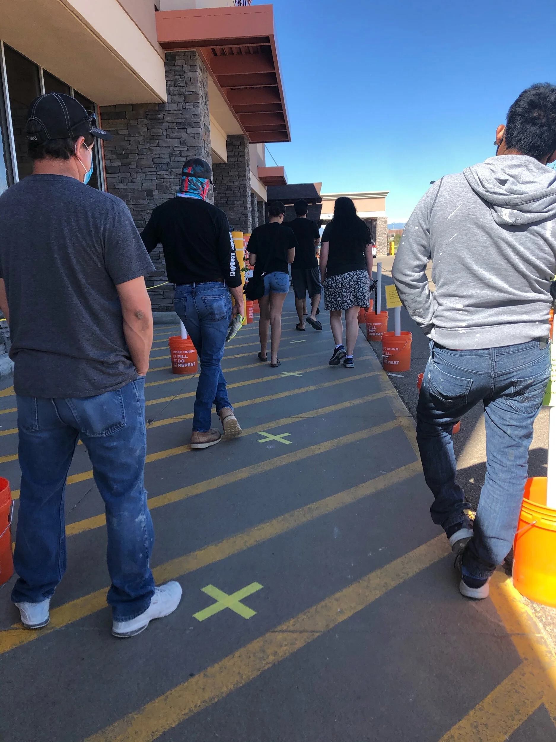 People lined up outside of a home improvement store, waiting to get their temperature taken before being allowed to enter the building. Remaining 6 feet apart.