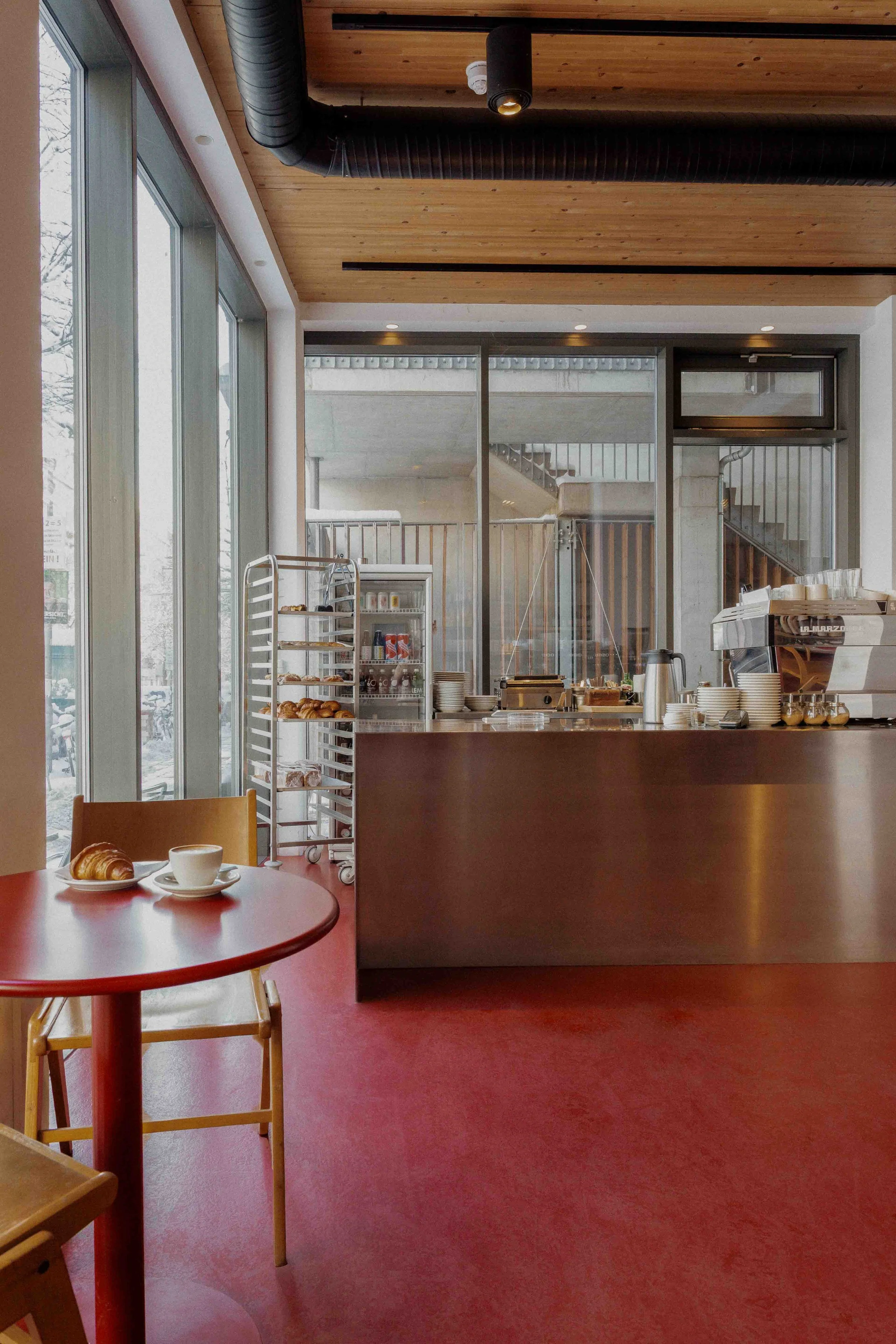 Open house in Pberg, Berlin: long steel counter beside a semi-matte oxblood-red floor with timber ceiling and metal shelving rack loaded with pastries.