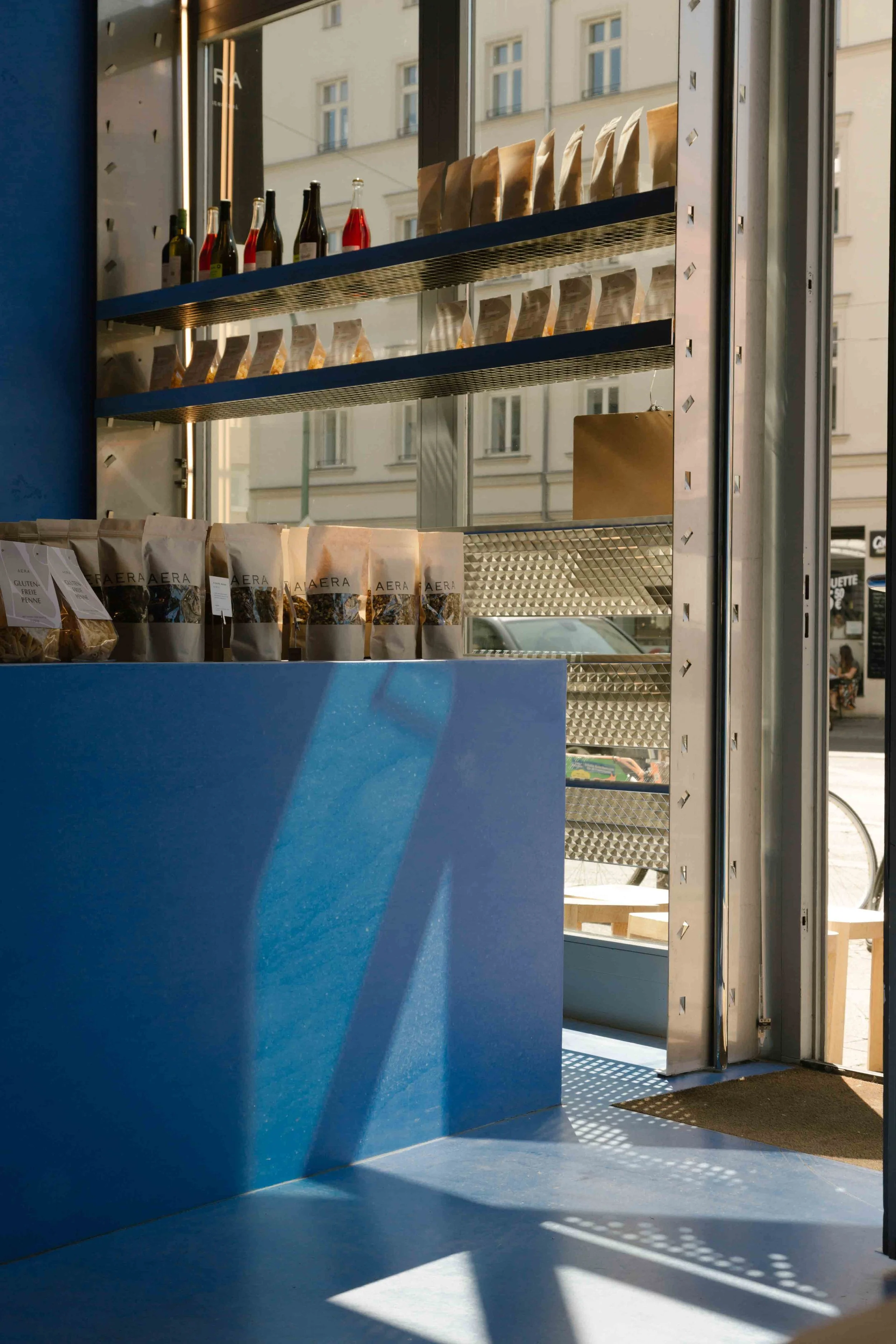 AERA Bread in charlottenburg, berlin: lapis lazuli blue concrete counter and shelving unit displaying packaged goods and glass bottles beside a glazed shopfront.
