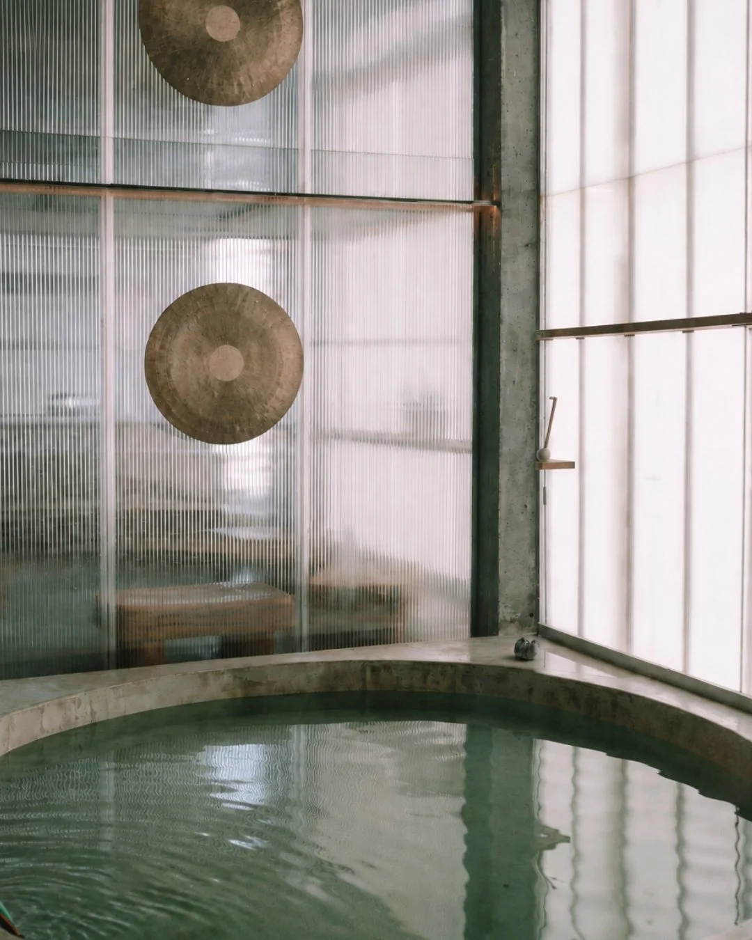 Circular plunge pool at ANTIspa Berlin, surrounded by translucent glass panels, exposed concrete and suspended metal gongs.