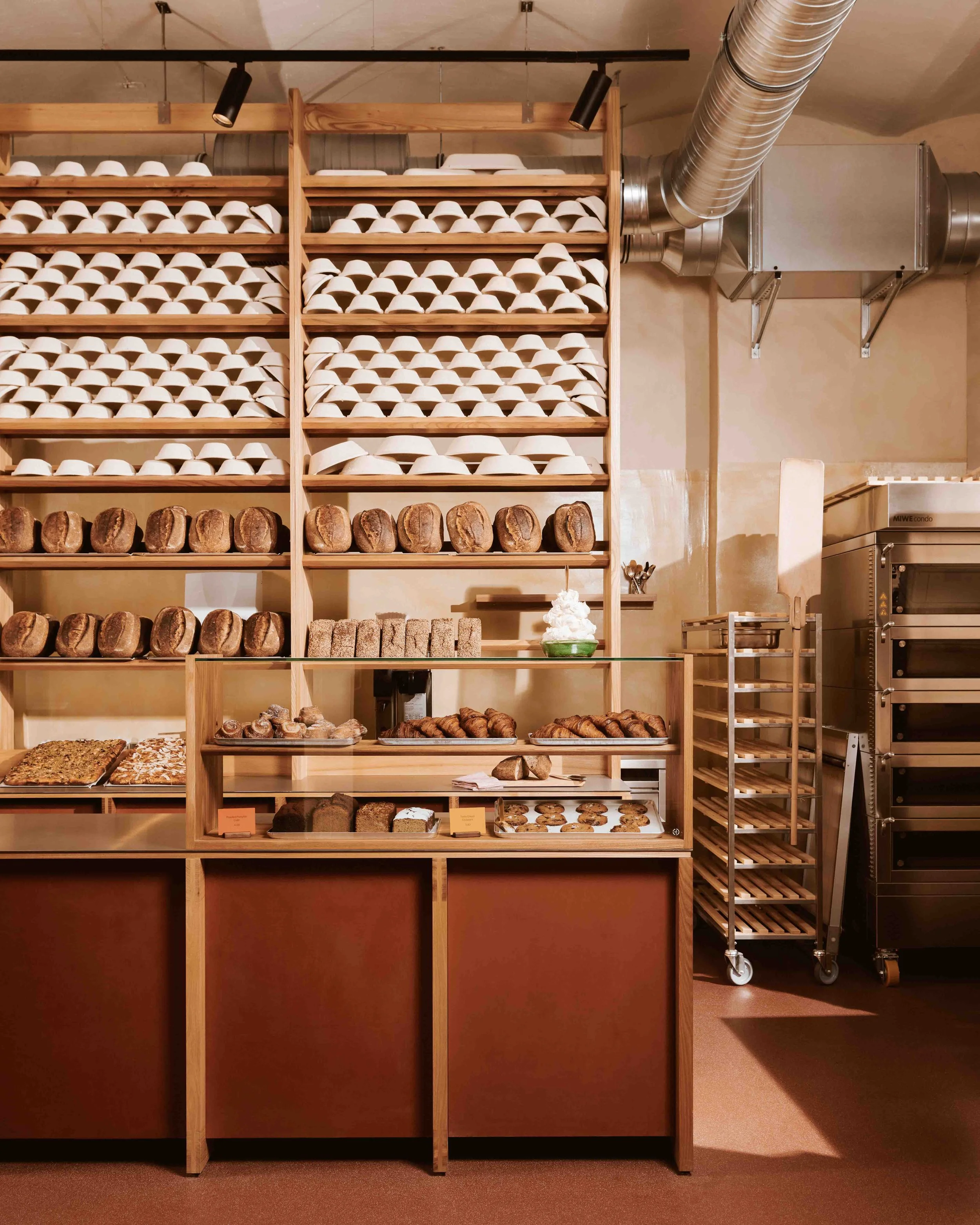 Timber shelving wall lined with proofing baskets and baked loaves above a glass display counter on a red vinyl floor