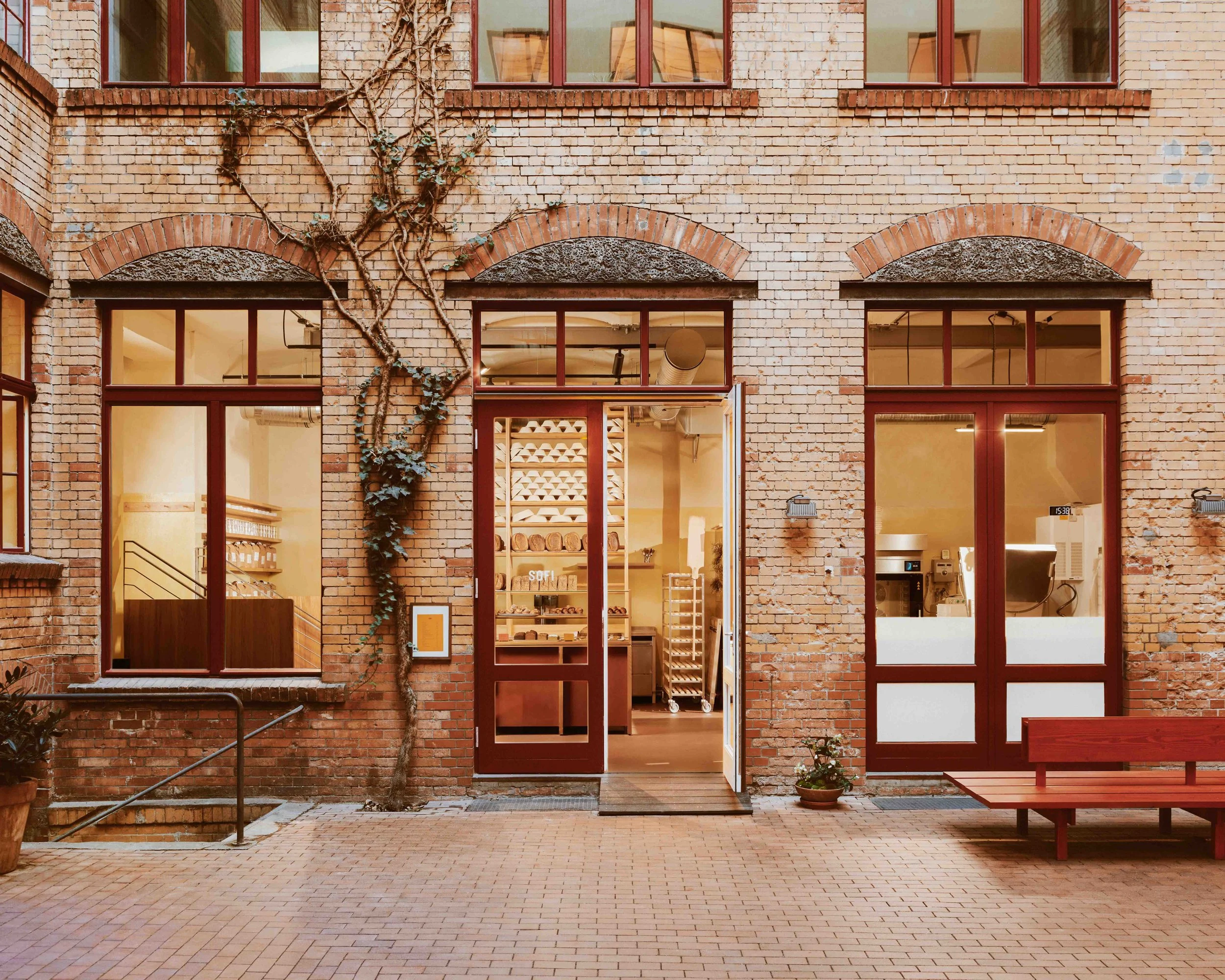 SOFI Mitte in Berlin: brick facade with arched transoms, dark-red timber windows and doors, open entrance revealing shelved interior, timber bench