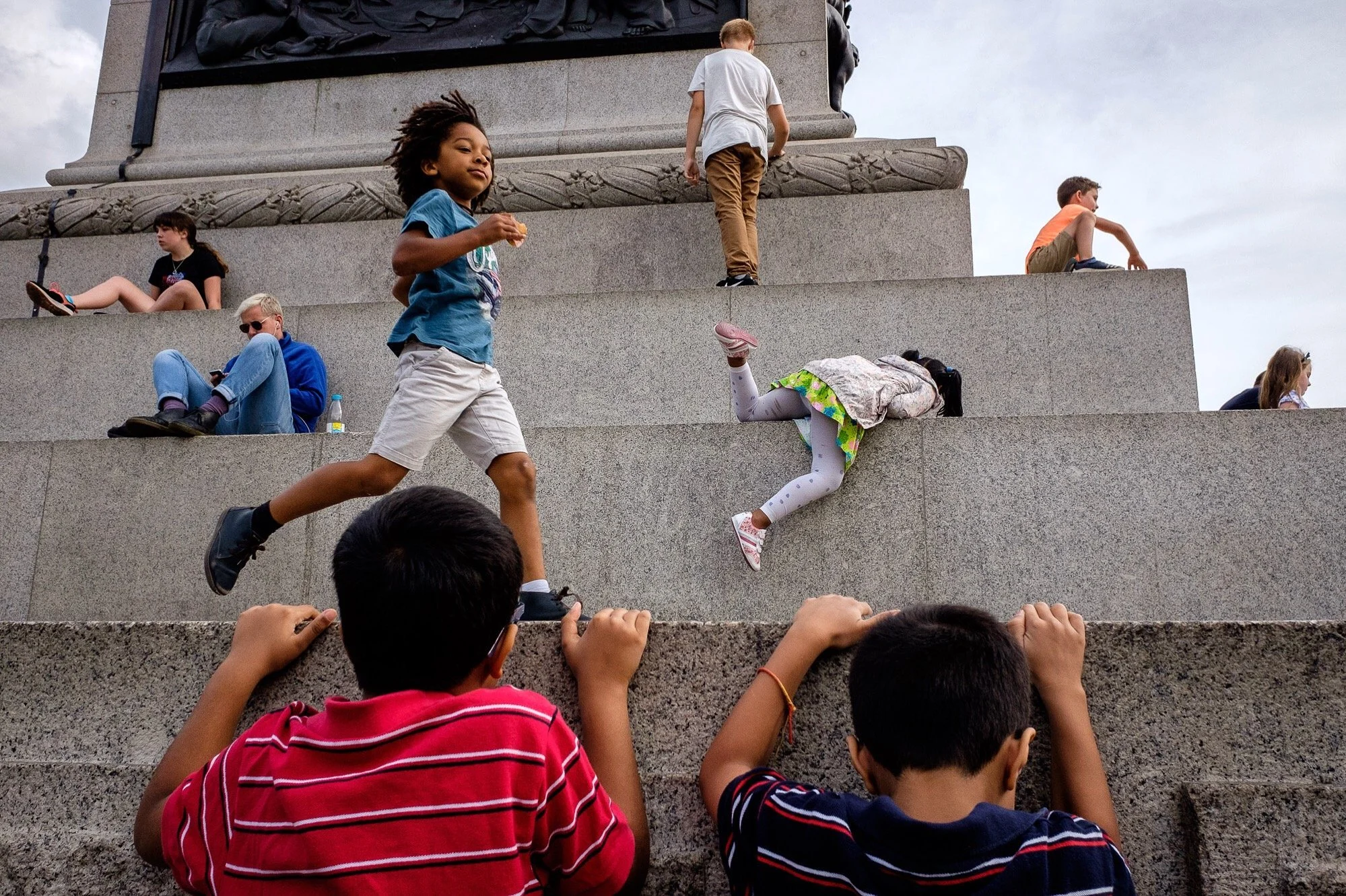  Children play on an outdoor stone staircase under an overcast sky. A boy in a blue shirt and white shorts runs up the stairs while a girl crawls with her head down a step below. Two boys in striped shirts are in the foreground with their backs to… 