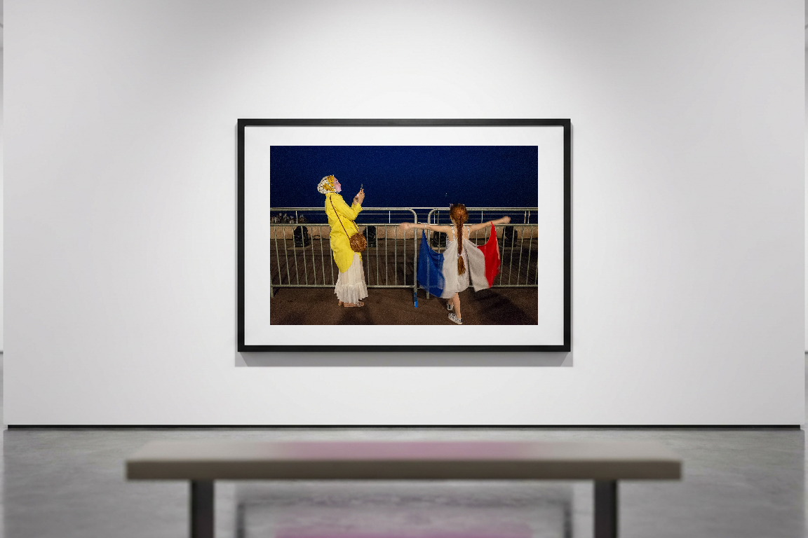 A woman in a yellow top and white skirt holds her phone up, looking at a girl in a white dress with red and blue fabric attached to her arms.…
