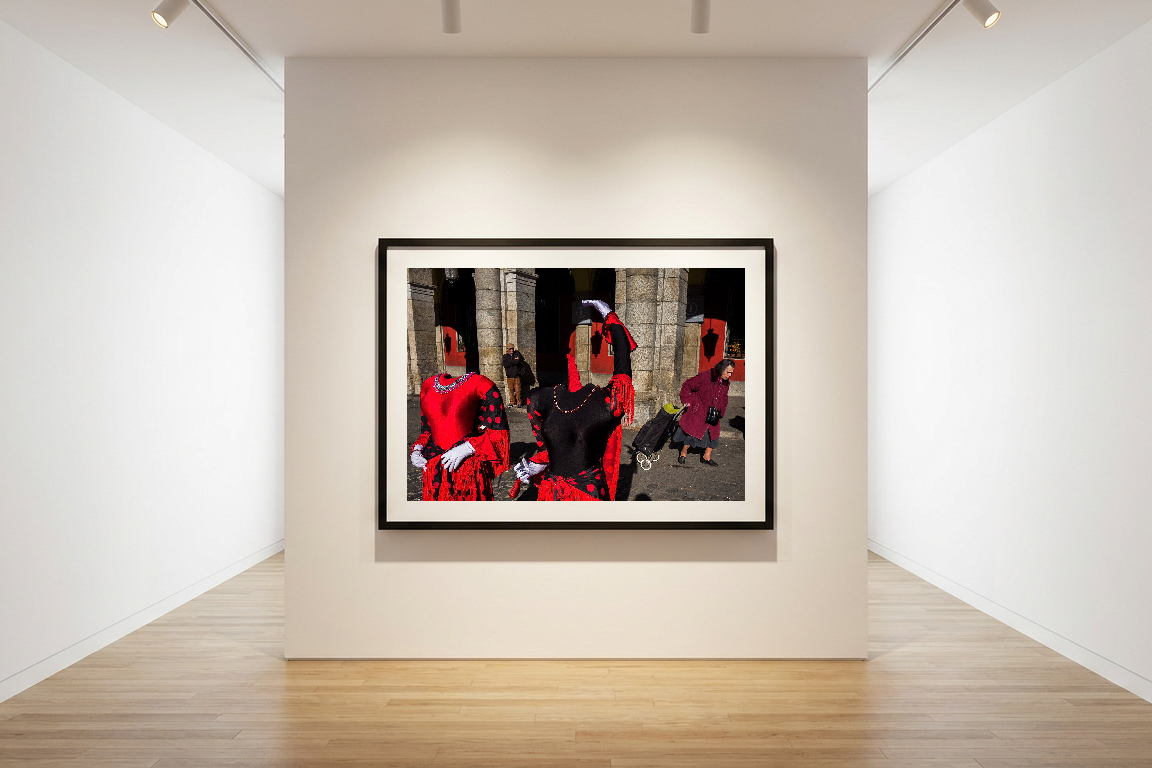 A framed photo of two headless mannequins in red dresses, mimicking flamenco dancers, in an outdoor setting. A person with dark hair walks by pushing a black cart.