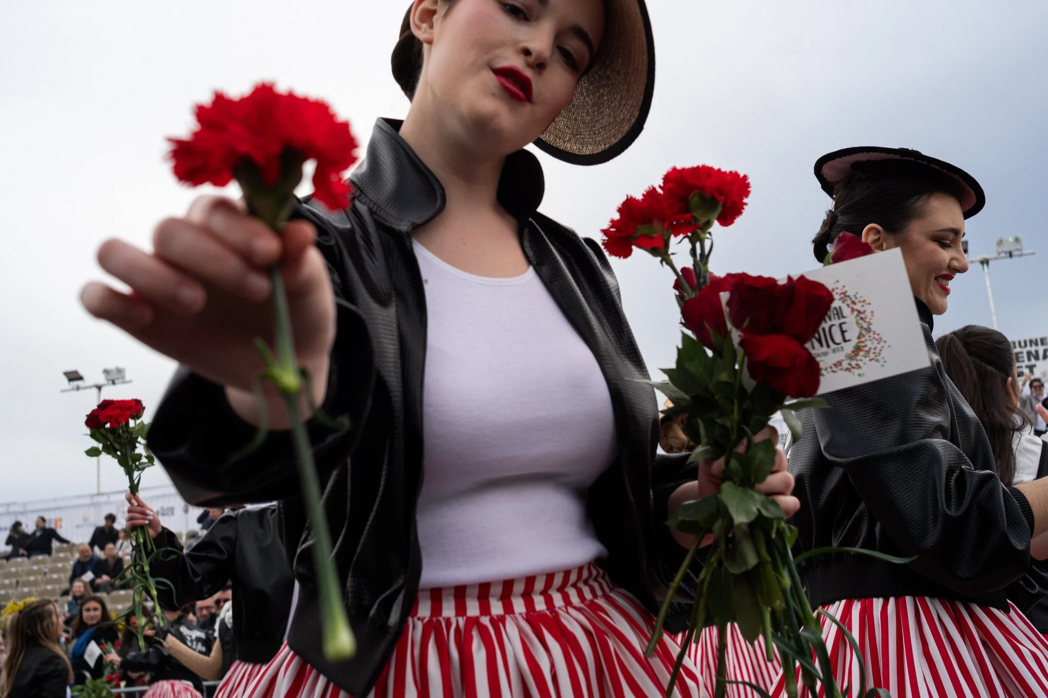 A woman in a black jacket and red and white striped skirt holds out a red carnation. She has dark hair, red lips, and wears a straw hat. Another woman is visible on the right holding red flowers.