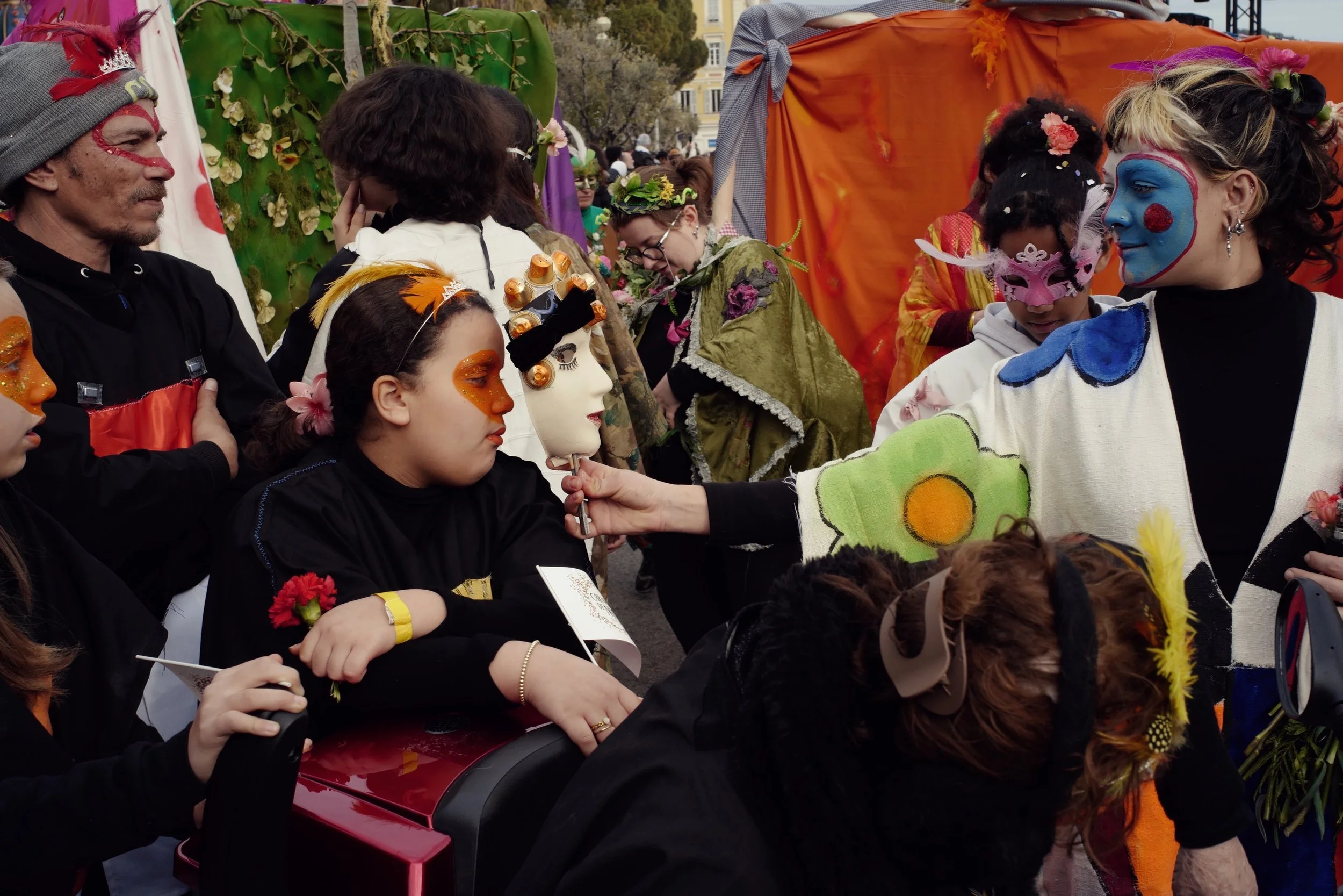 A man with red face paint and a feathered beanie sits in a crowd. Another person with a yellow headband holds a mannequin head. Other people in costumes and face paint are nearby.